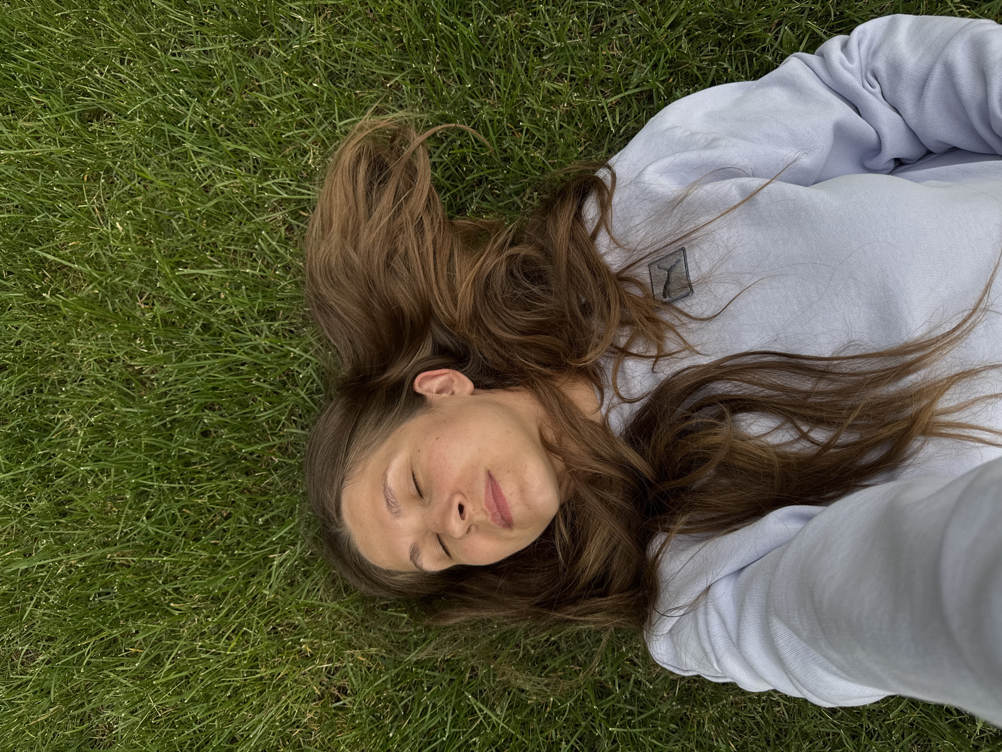 A young woman is lying on her back on a lawn. Her eyes are closed and her head is slightly tilted. Her long, brown hair is spread out around her head. She is wearing a light blue sweatshirt with a Puma logo on the left chest. The lawn is a vibrant green and appears to be well-maintained. The image is taken from a high angle, looking down at the woman. There is no indication of the specific location within Gatineau, Canada.