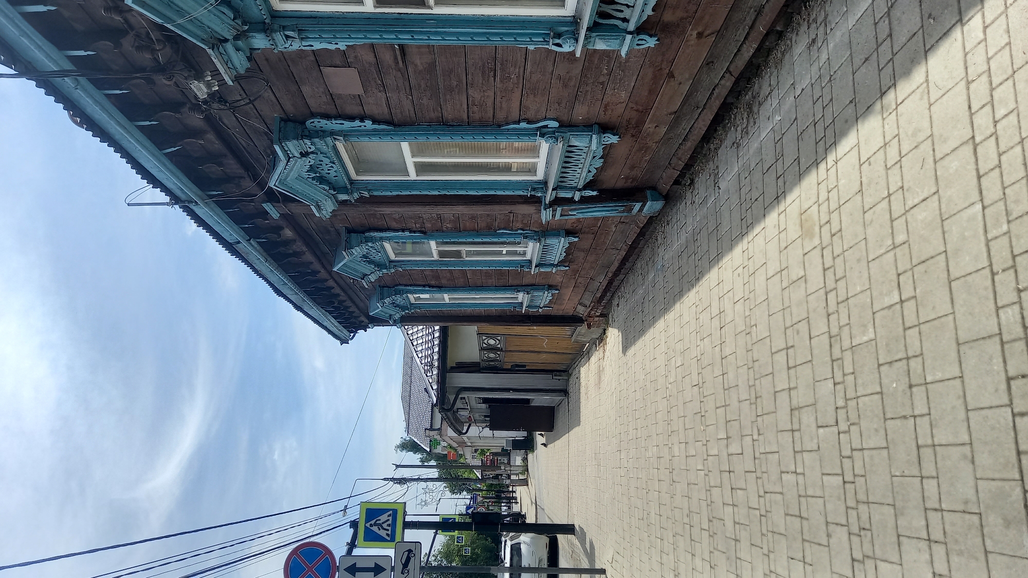 The image displays a street scene in Zvenigorod, Russia, captured during daylight under a partly cloudy sky. The perspective is from a low angle, looking along a light grey paved sidewalk.
To the immediate right, a multi-story wooden building constructed of dark brown horizontal planks occupies a significant portion of the frame. This structure features prominent, intricately carved light blue wooden window frames (nalichniki) around its white-framed windows. Four complete window sections are visible, along with a partial fifth on the far right. A decorative, carved light blue frieze runs along the building's dark corrugated metal roofline, which is partially visible along with a light blue gutter. Electrical wires and a utility meter are attached to the building's facade. A strong diagonal shadow from the building extends across the sidewalk.
The sidewalk in the foreground is composed of interlocking light grey concrete pavers. Two pigeons are situated on this sidewalk in the middle ground, near a utility pole.
On the left side of the frame, a series of utility poles supports numerous black overhead wires. Mounted on these poles are several traffic signs: a red circular sign with a blue background and a red diagonal cross, typically indicating "No Stopping" or "No Parking"; a white rectangular sign with vertical up and down arrows; a blue square sign featuring a white pedestrian icon, indicating a pedestrian crossing; and a blue square sign with a white cargo truck icon.
Further down the street on the left, a white car is parked parallel to the curb. Beyond this, additional buildings are visible, including one with a dark tiled roof and another with a dark corrugated roof and lighter walls. A wooden fence or gate is present between the ornate wooden building and the subsequent structure. Green foliage from trees is visible in the background.
The sky is a light blue with scattered, wispy white clouds. No human figures are present within the observable scene.