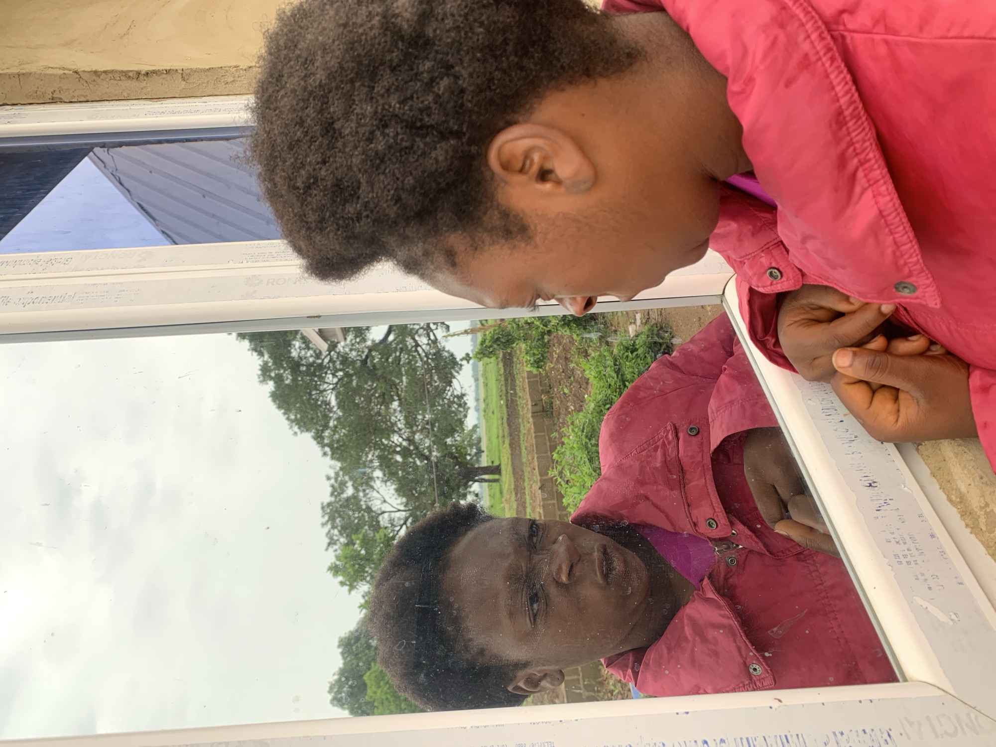 A person with dark skin and short, curly black hair, wearing a red jacket, is reflected in a window. The reflection shows the person's face clearly. Outside the window, a rural landscape is visible, including green fields, trees, and a cloudy sky. The window frame is white. The location is Agban, Nigeria.