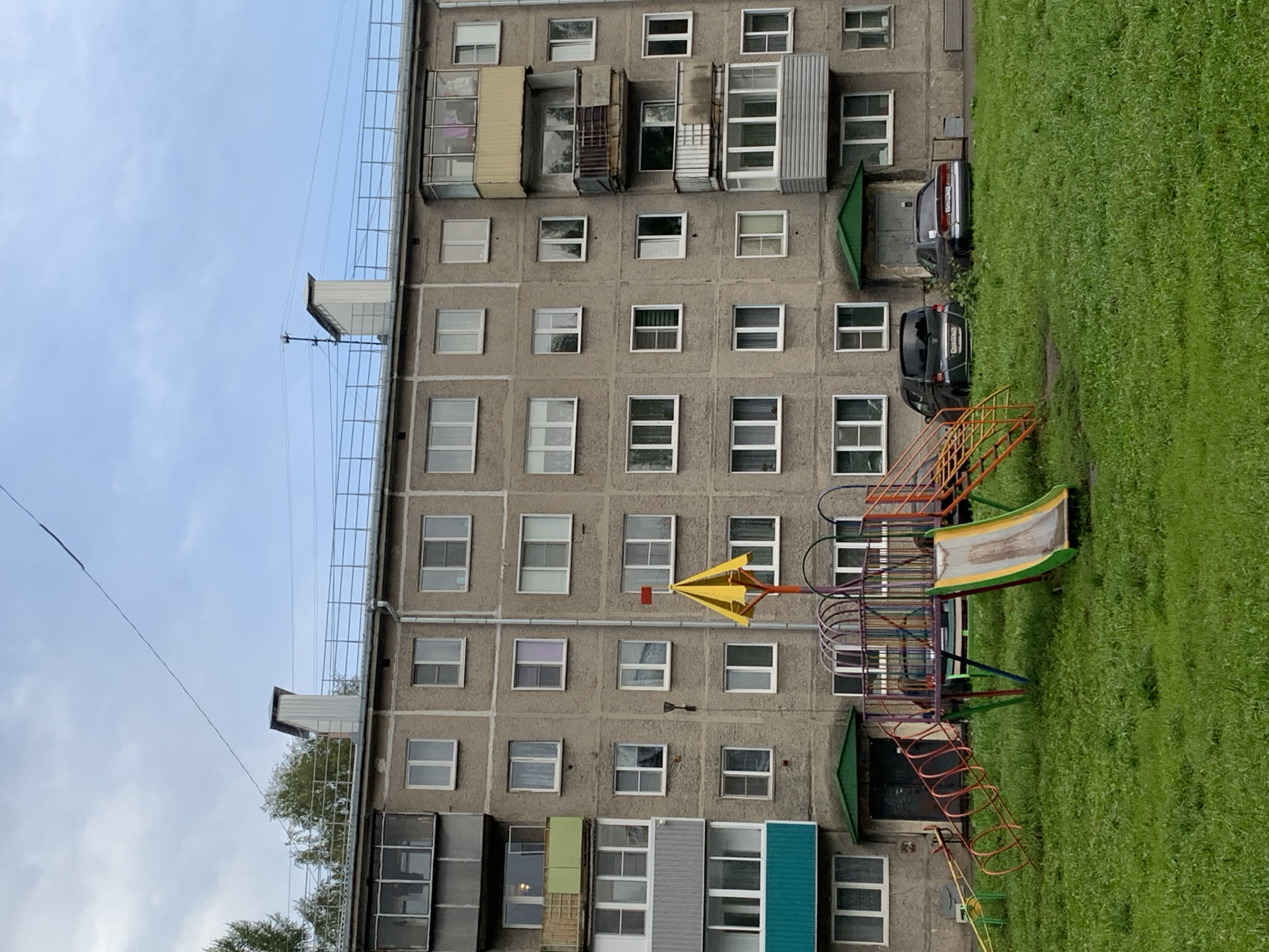 The image shows a concrete apartment building in Prokopyevsk, Russia, with a playground in front of it. The building is several stories tall and has numerous windows. There are balconies on some of the floors. The playground features a slide, a climbing structure, and a seesaw. Two cars are parked on the grass in front of the building. The sky is cloudy. The overall scene depicts a typical urban residential area.
