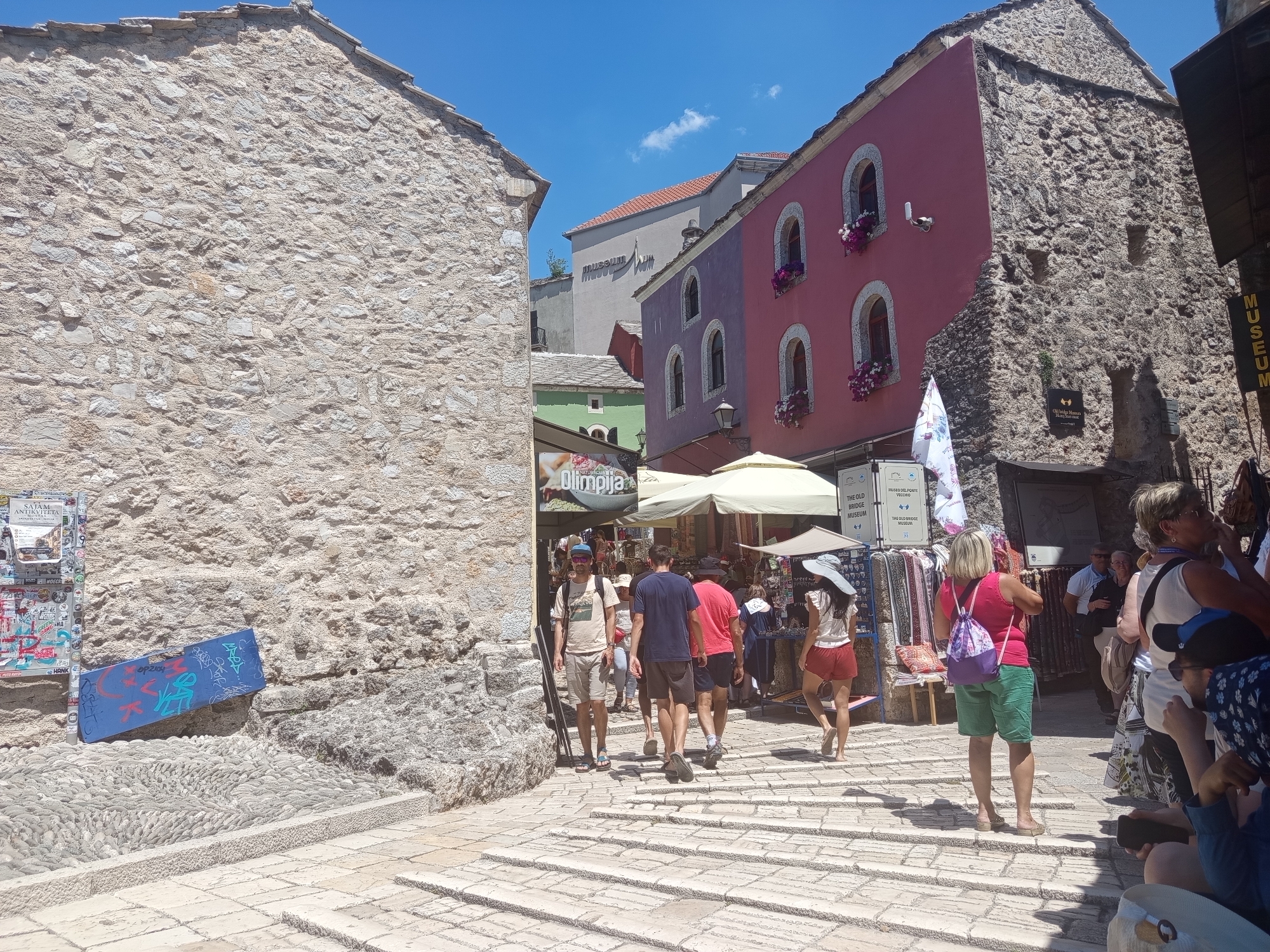 This media file captures a bustling scene in the historic old town of Mostar, Bosnia and Herzegovina. The image is dominated by the distinctive stone architecture of the region, with a prominent rough-hewn stone building on the left and colorful, multi-story buildings on the right, featuring pink and purple facades with arched windows adorned with flowers. A bright blue sky with scattered white clouds suggests a sunny day, likely mid-morning or early afternoon, given the strong shadows.
Several people are visible, appearing to be tourists, navigating a stone-paved walkway that slopes upwards. They are dressed in casual summer clothing, indicative of warm weather. Some are walking away from the viewer, while others are facing towards the camera. In the background, market stalls with awnings are set up, displaying goods and adding to the lively atmosphere. Signs in the image include "MUSEUM," "SAJAM ANTIKVITETA" (Antique Fair), and "Olimpija," suggesting commercial activity and cultural heritage sites. Another sign reads "THE OLD BRIDGE MUSEUM," a key landmark in Mostar. A blue board with graffiti is leaning against a stone wall on the left, adding a touch of urban art to the ancient setting. The overall impression is one of a vibrant tourist destination, showcasing the blend of historical architecture, local culture, and contemporary life on a beautiful sunny day.