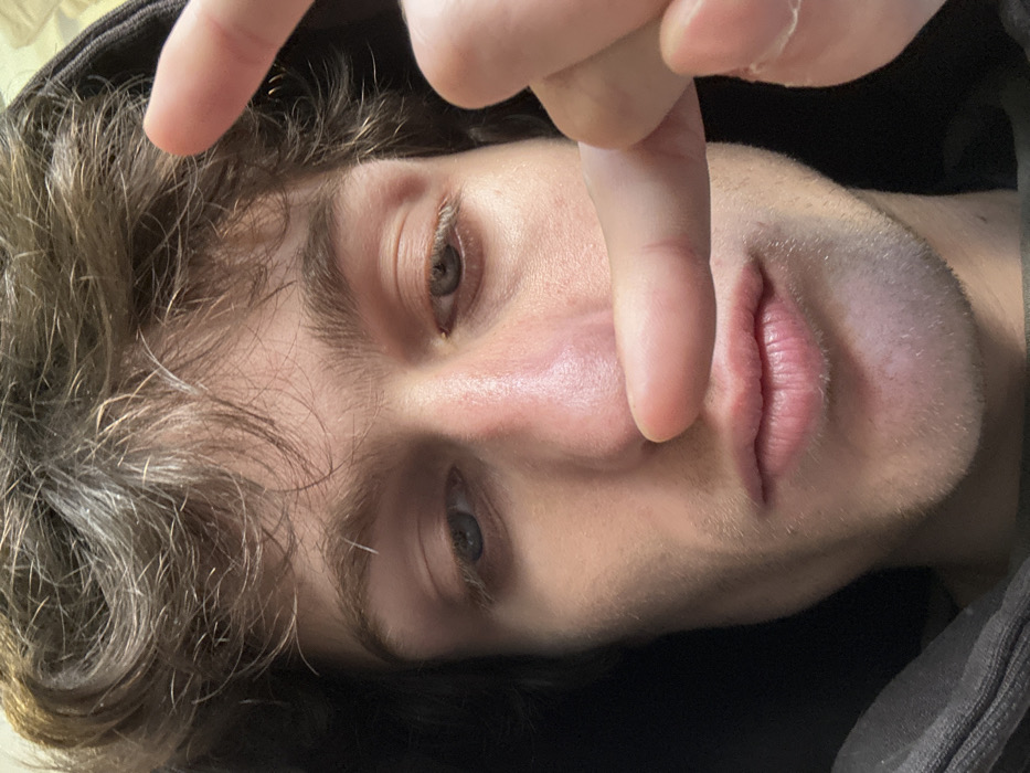 A close-up, frontal portrait captures a male individual with light skin, disheveled brown hair covering his forehead, and prominent brown eyebrows. His left eye appears blue-grey, while his right eye is a lighter brown or amber hue. A light stubble is visible on his chin and upper lip. He is wearing a dark, possibly hooded, garment. His right index finger is positioned horizontally across his mid-face, obscuring his philtrum and upper lip, touching the base of his nose. A portion of another finger, likely the right thumb or middle finger, is visible on the far right of the frame, near his temple. The background is uniformly dark and out of focus, providing no discernible environmental details. The lighting illuminates his face from the front. The scene is situated in Vincennes, France.