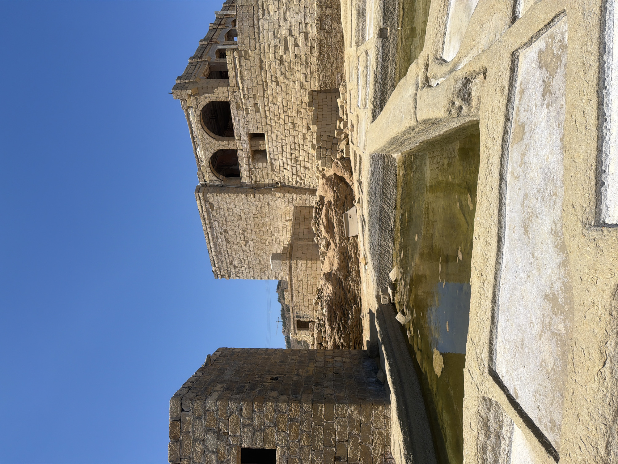 This is a vertical, eye-level shot of the Salt Pans in Marsalforn, Malta, under a clear blue sky. In the foreground, there are shallow, rectangular pools carved into the rock, some filled with murky, green water and others dry and encrusted with white salt. A thick, carved stone barrier separates the pools. In the midground, a weathered, rectangular stone building with a dark window opening stands to the left. To the right of it, the ruins of a larger, multi-story stone structure rise up, featuring arched openings on its upper levels and a complex, uneven stone facade. The structures appear ancient and in a state of disrepair, with rubble and displaced stones scattered around their bases. The overall environment is arid and sun-drenched.