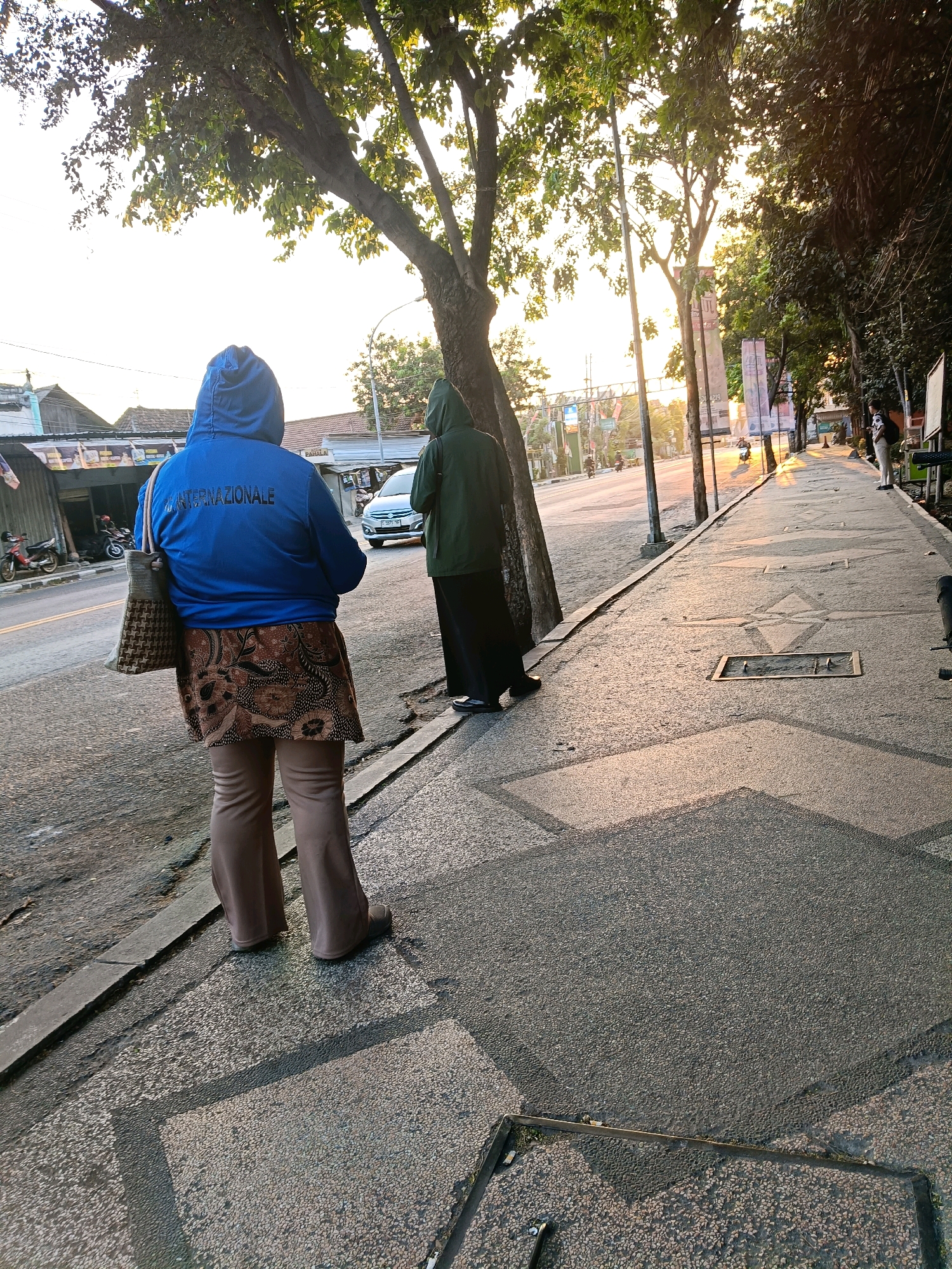 An outdoor, eye-level, medium shot captures a street scene at sunset in Lamongan, Indonesia, with two individuals visible. On the left, a person wearing a blue hooded jacket with "INTERNAZIONALE" printed in white on the back and a brown patterned skirt over beige flared pants, carries a woven bag. To their right, another person in a green hooded jacket and black pants stands facing away from the camera. Both are on a sidewalk next to a road where a silver car is driving. The setting sun casts long shadows and illuminates the trees lining the street and the patterned sidewalk. The background shows buildings, streetlights, and more trees, suggesting a moderately urban environment.