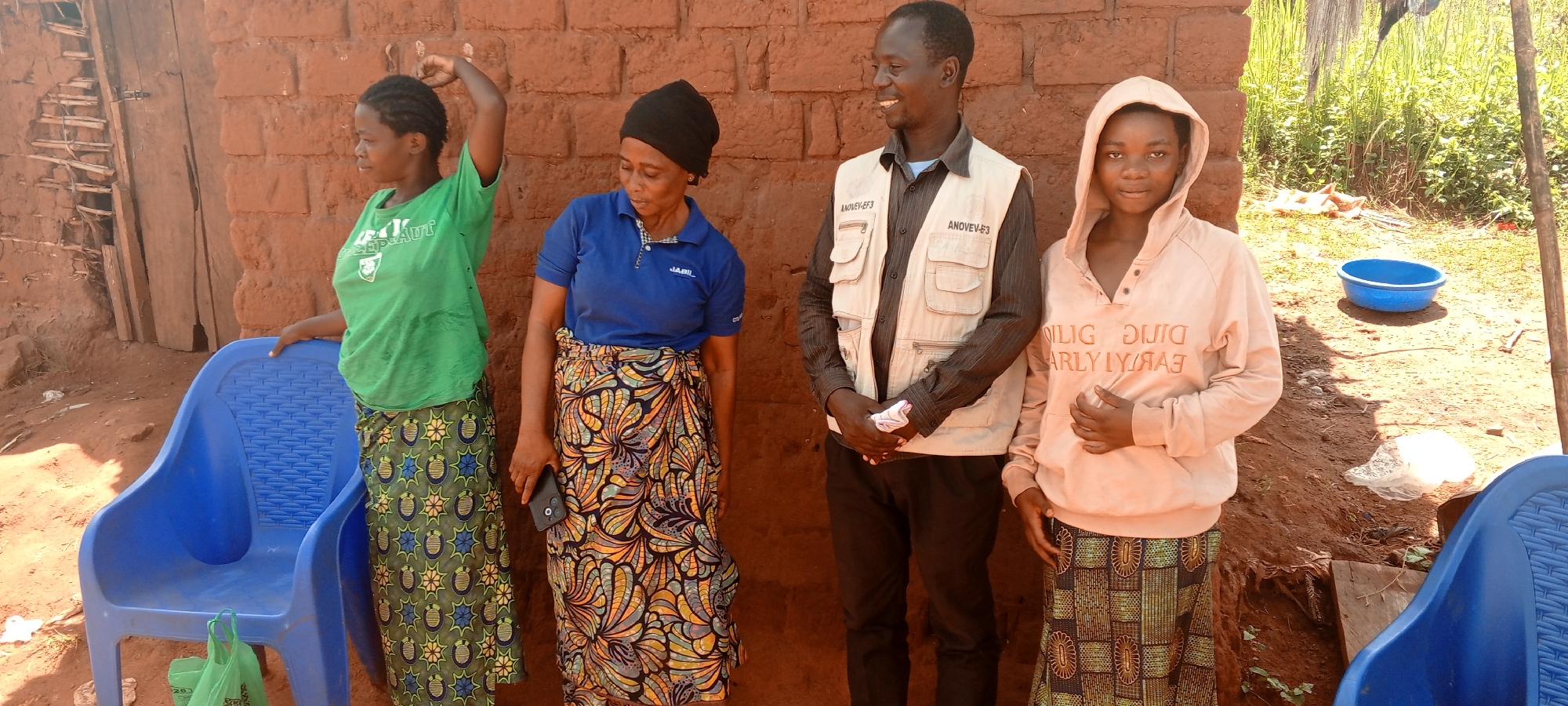 Four individuals, three women and one man, are standing outdoors in Makungu, Democratic Republic of the Congo, in bright daylight. They are positioned in front of a reddish-brown brick or mud-brick wall.
On the far left, a woman with braided hair wears a green t-shirt and a green-patterned skirt, her left arm raised. She leans against a blue plastic chair, at the base of which a green plastic bag rests. Next to her, a woman in a blue polo shirt, a black headscarf, and a vibrant, multi-colored patterned skirt holds a black smartphone. The text "JABIL" is visible on her polo shirt.
To their right, a man in a dark shirt and a light-colored utility vest smiles, looking to his left. His vest has "ANOVEV-EF3" printed on both chest pockets. On the far right, a young woman wears a peach-colored hooded sweatshirt with stylized and partially reversed text that appears to read "DILIG DUIG EARLY LYRAA" (likely "DILIGENT EARLY RAISER") and a patterned skirt. She looks directly at the viewer.
The ground is dirt, and in the background on the right, there is a blue plastic bowl and lush green vegetation. The overall setting suggests a casual gathering in a rural or semi-urban environment.