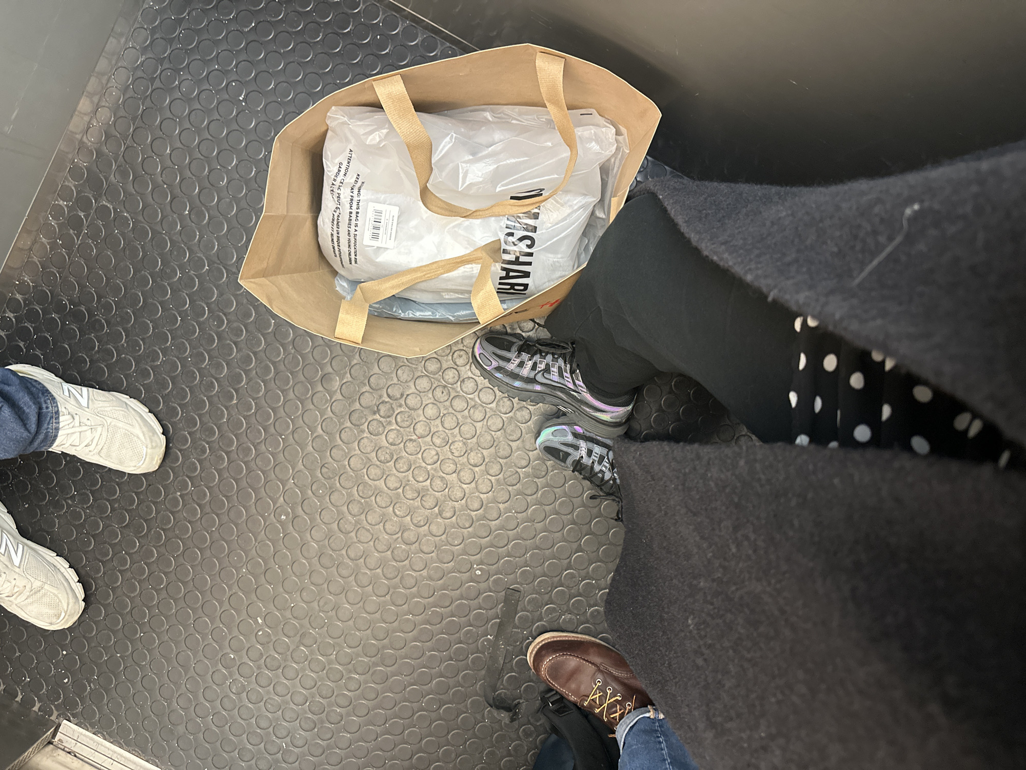 The image depicts a low-angle view of the feet of at least three individuals and a shopping bag on a dark grey or black rubberized floor featuring a regular circular stud pattern. This flooring is characteristic of London's public transport, specifically the London Underground or a London bus.
In the immediate foreground, a person's lower legs are visible, clothed in dark trousers or leggings, partially obscured by a dark, possibly wool, coat. White polka-dotted fabric is visible beneath the coat on the right leg. The person wears dark athletic shoes with iridescent purple/blue accents and black laces. A light brown paper shopping bag with flat fabric handles sits on the floor to the right of this person's feet. Inside the bag, a clear plastic garment bag containing white clothing items is visible. The word "GYMSHARK" is printed in black on the white clothing, and a warning label in multiple languages is present on the plastic bag, including "WARNING! THIS BAG IS A SUFFOCATION RISK."
To the left of the foreground individual, another person's lower leg, clad in blue denim jeans, is visible, wearing a brown leather lace-up boot with light brown laces and distinct moc-toe stitching. In the upper portion of the frame, a third person's feet are visible, wearing blue denim jeans and light grey New Balance athletic sneakers, with the "N" logo clearly visible on one shoe. The rubberized floor shows signs of minor wear and scattered white specks. The scene captures passengers in a confined public transport environment in the city of London, United Kingdom.