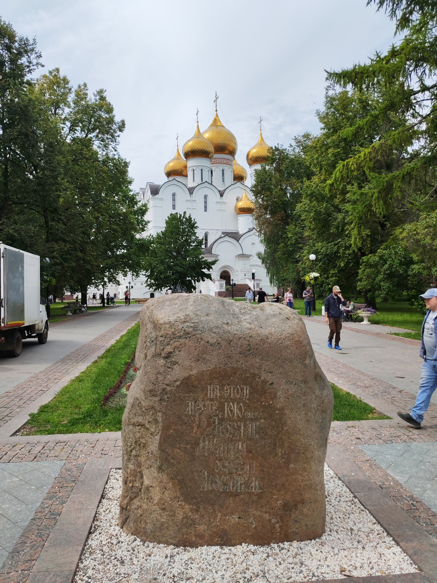 A large, roughly textured, reddish-brown boulder with carved inscriptions is positioned in the foreground of the image. The inscriptions are in Cyrillic script and appear to be weathered. The boulder rests on a bed of small white pebbles. Behind the boulder, a paved pathway leads towards a white building with multiple golden domes, characteristic of an Orthodox church. Lush green trees with full foliage flank the pathway and surround the building. Several people are visible walking on the pathway and sitting on benches in the background, some near the church entrance. A white truck is parked on the left side of the pathway. The sky is overcast. The location is identified as Rusanovo, Russia.
