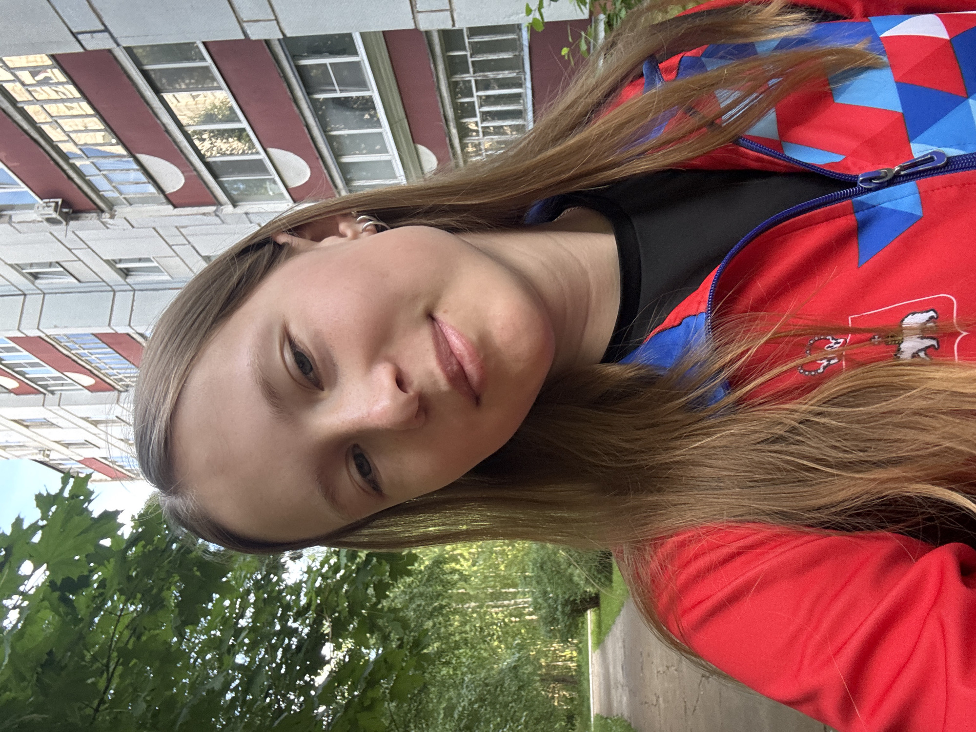 A close-up, slightly tilted portrait photograph, oriented 90 degrees counter-clockwise. The subject is a young person with long, light brown hair, appearing to be female, captured in an outdoor setting during the daytime. She is looking slightly upwards towards the camera with a subtle smile or smirk. She wears small silver hoop earrings.
She is dressed in a red jacket featuring a geometric pattern of blue and white triangles and squares, with a visible blue zipper. A black top is visible underneath. On the right side of the jacket, partially obscured by her hair, there is a white emblem or logo that appears to include a stylized figure and some indistinct Cyrillic characters.
The background consists of a multi-story building to the left, which has a light-colored (white or grey) facade with distinct horizontal red bands and numerous rectangular windows, some reflecting the sky or sunlight. To the right and below the subject, lush green trees and foliage are visible, suggesting a park or a treelined area. A paved path is faintly visible through the trees in the lower right background. The bright, natural lighting indicates it is daytime, likely sunny or partly sunny.
The setting is an urban or suburban outdoor environment, specifically Chaikovsky, Russia, with architecture typical of the region and abundant greenery.