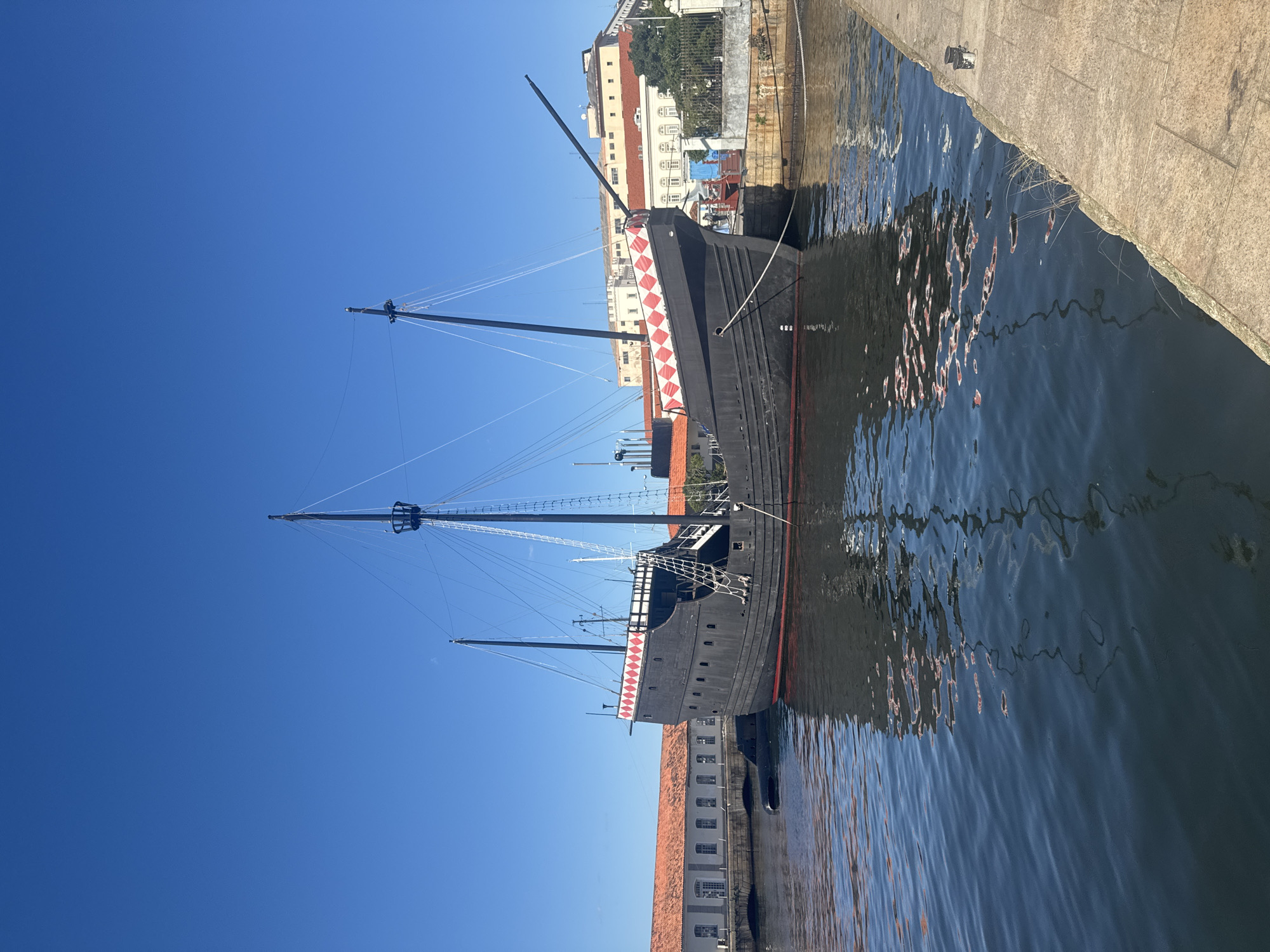 A historical replica of a caravel ship is docked in a canal in Rio de Janeiro, Brazil. The ship is painted black with a red stripe along the waterline and a decorative band of red and white diamonds near the bow. It has two masts with rigging and a crow's nest on the taller mast. The ship is moored alongside a stone quay, and reflections of the ship and surrounding buildings are visible on the calm water. In the background, there are several low-rise buildings with orange tile roofs and white walls. The sky is a clear, vibrant blue.