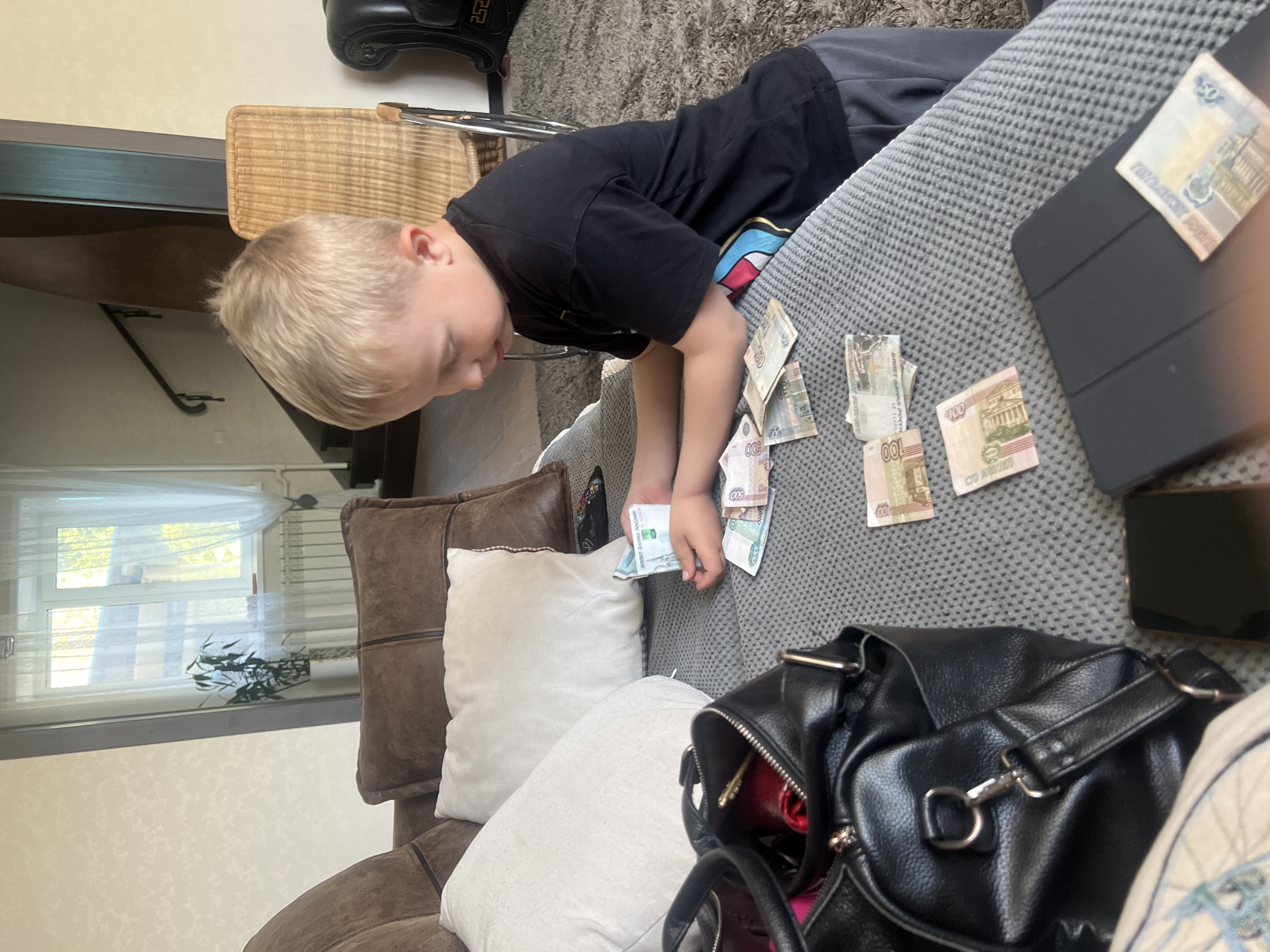 A boy with blond hair wearing a black t-shirt and gray pants leans over a gray couch, holding several Russian rubles in his hand. Scattered on the couch are other Russian rubles, a black tablet with a cover, and a black smartphone. A black leather handbag sits on the couch near the boy. Behind the couch are pillows and a window with white curtains. In the background, there is a wicker chair and part of a staircase. The location is Unknown, Unknown.