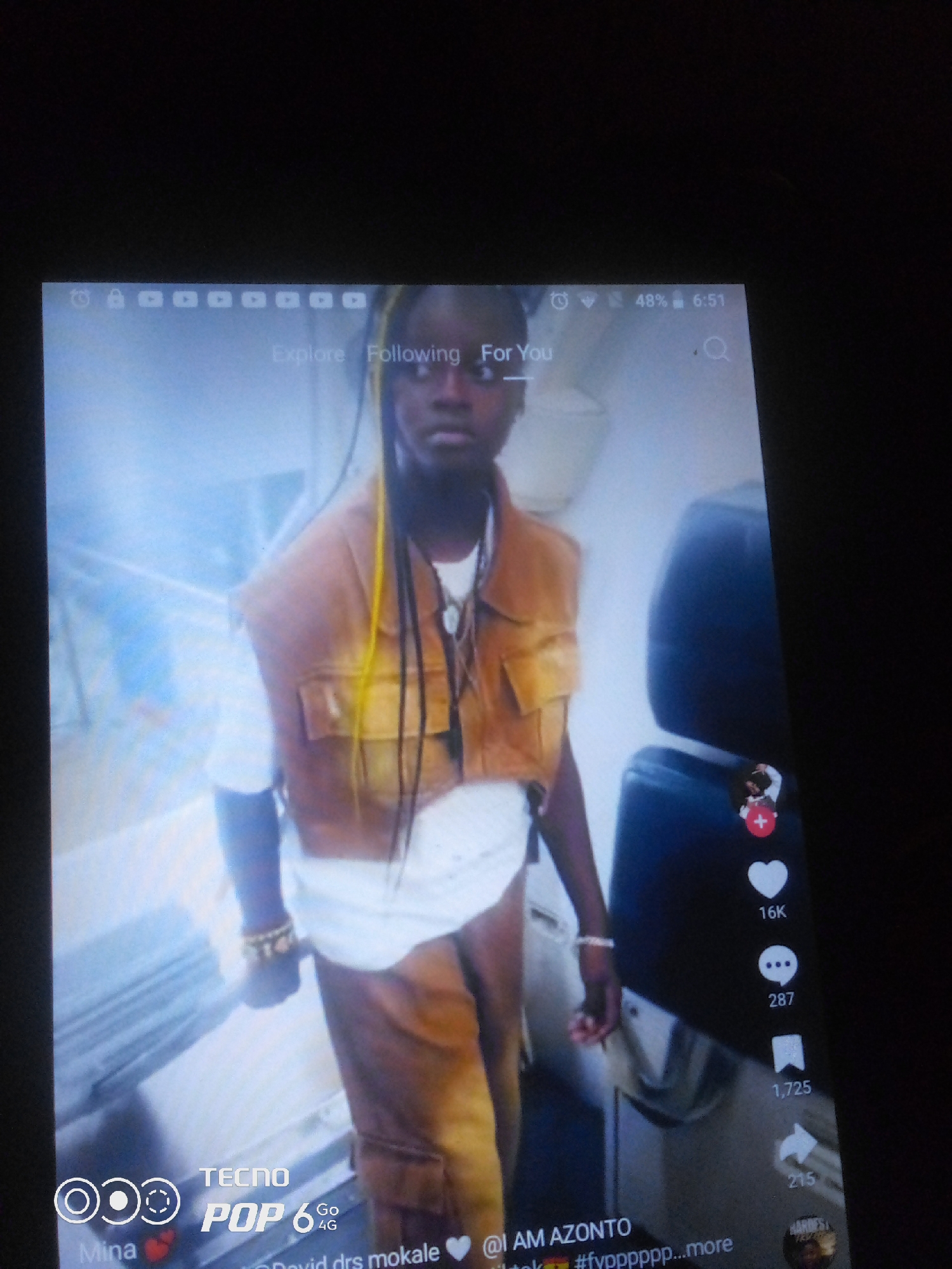 A person with dark skin and yellow and black braided hair is standing in front of a blurry background. They are wearing a brown vest over a white t-shirt and brown pants. There are several icons on the right side of the image, including a heart with the number 16K next to it, a speech bubble with the number 287, and a folded paper with the number 1,725. At the bottom of the image, there is text that reads "TECNO POP 6 Go 4G". There is no indication of the location being in Howayi, Nigeria or any other specific city.