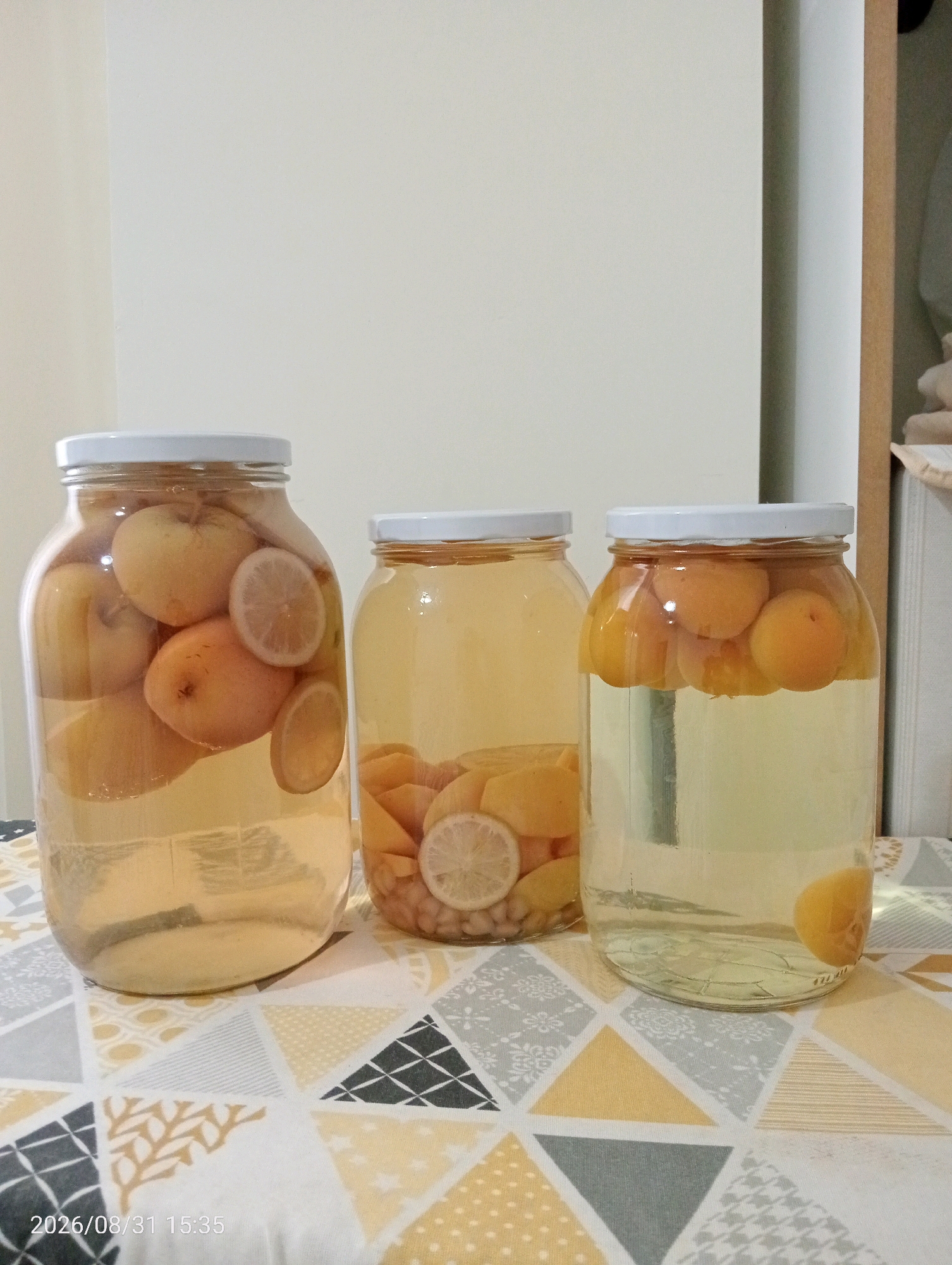 Three glass jars filled with fruit and liquid are arranged on a patterned tablecloth. The jar on the left contains whole apples and lemon slices. The middle jar contains what appears to be peaches or apricots cut into wedges, along with some round, light-colored objects. The jar on the right holds whole apricots. The background is a plain, light-colored wall. A timestamp in the bottom left corner reads "2026/08/31 15:35". The image was taken in Silifke, Turkey.