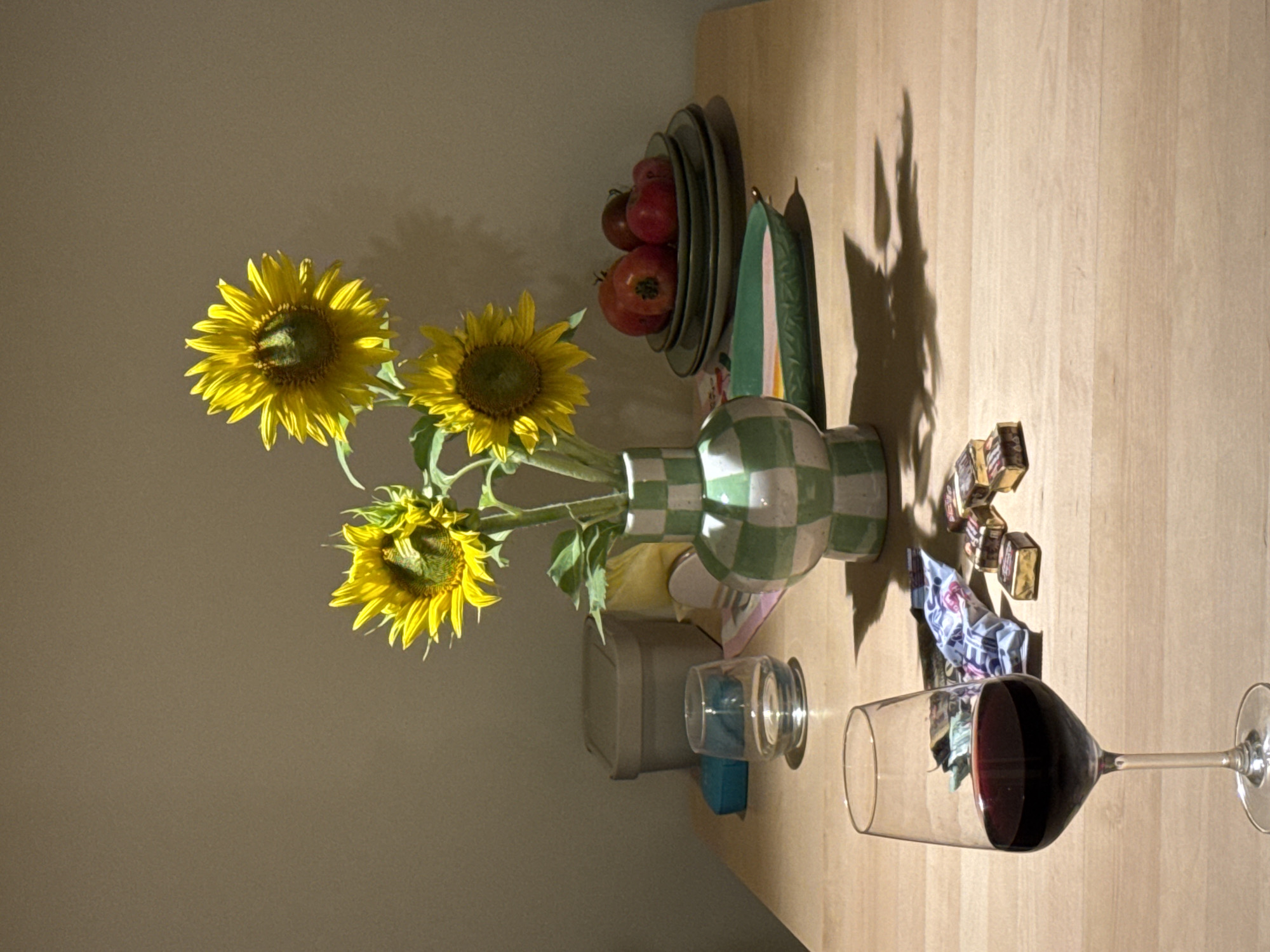 The image depicts a still life scene on a wooden table. In the foreground, a wine glass filled with red wine is positioned to the left. To the right of the wine glass, there are several individually wrapped chocolates and a crumpled piece of patterned fabric.
Behind the wine glass and chocolates, a vase with a green and white checkered pattern holds three sunflowers. To the left of the vase, there is a clear glass with a small amount of liquid. Further back on the table, there is a beige storage container.
To the right of the vase, a stack of dark green plates holds several red fruits, likely apples or tomatoes. In front of the plates is a long, shallow green dish. The background is a plain, light-colored wall, and the scene is illuminated by a light source casting shadows on the table and wall.
The image does not contain any elements that would allow for specific location identification within the city of Izhevsk, Russia.