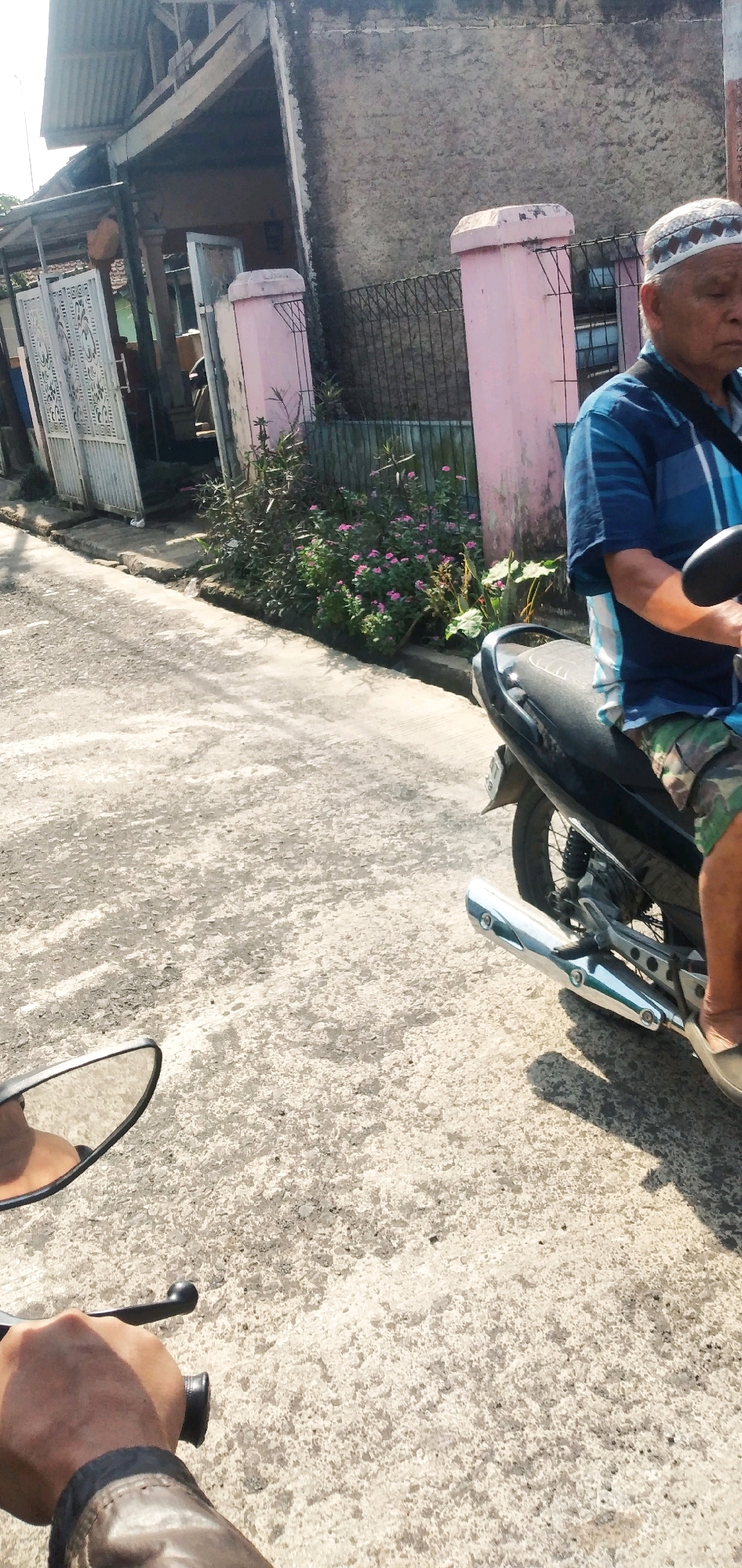 An elderly man wearing a white skullcap and a blue and white striped shirt is riding a black motorcycle on a paved road in Cilaku, Indonesia. He is wearing camouflage shorts and tan shoes. The man is looking down and to the right. In the background, there is a building with a grey concrete wall and a corrugated metal roof. There is also a pink pillar and a black metal fence. A bush with pink flowers is growing near the fence. The road is cracked and weathered. The image appears to be taken from another motorcycle, as a hand holding a handlebar is visible in the lower left corner, and a rearview mirror is also visible. The sun is shining brightly, casting shadows on the road.