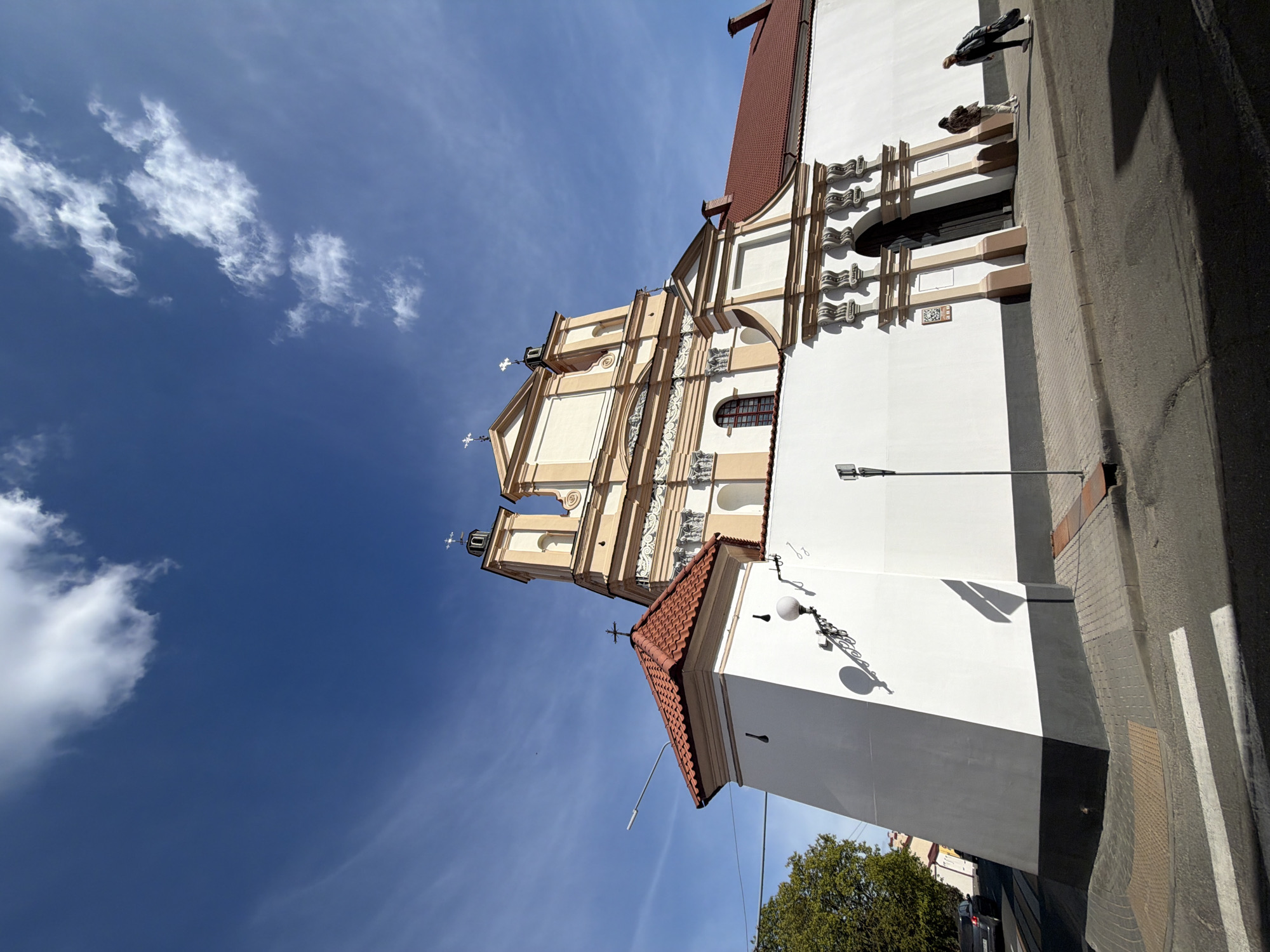The image, oriented vertically, showcases a grand, ornate church building under a bright blue sky in Hrodna, Belarus. The main structure features a multi-tiered facade in cream and white hues, adorned with intricate baroque architectural details, including decorative columns, arched windows, and cornices. Two prominent bell towers, each topped with a cross, rise towards the sky. Sections of the church's roof are covered with reddish-brown tiles.
The sky is a clear, deep blue, with scattered white, wispy clouds. Bright sunlight illuminates the scene, casting distinct shadows from the building and other elements, indicating a sunny day, likely midday or early afternoon.
On the right side of the image, two individuals are visible, walking or standing on a paved sidewalk or road that runs alongside the church. A modern lamppost is present in the lower right foreground, and a small, mature tree with green foliage is visible at the bottom left. A small, unreadable plaque is affixed to the lower white wall of the church. The overall atmosphere is peaceful, highlighting the historical architecture against a vibrant natural backdrop.