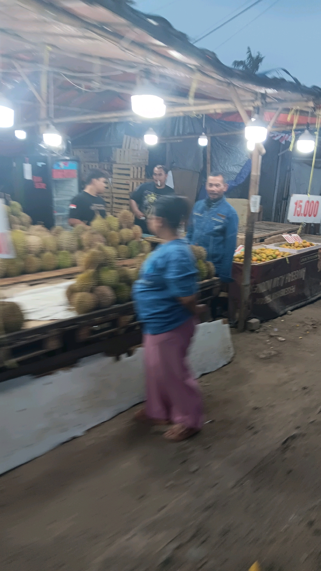 This is a medium shot, slightly blurred, outdoor image taken at a fruit stall, likely in Depok, Indonesia, as indicated by the provided context. The scene is illuminated by bright overhead lights, suggesting it is evening or nighttime. In the foreground, a person in a blue shirt and pink pants is walking away from the camera, their legs blurred by motion. To the left, there is a large display of durian fruit, with some being sold from wooden crates. Several people are visible behind the durian stall; one man in a black t-shirt with an Indonesian flag emblem is looking towards the camera, while another man in a black t-shirt is visible to his right. Further to the right, a man in a blue denim jacket is standing, looking towards the durian stall. Behind him, a sign displays "15.000" in red digits, likely indicating a price. In the far right of the frame, another stall displays bright orange fruits, possibly tangerines or oranges, also in wooden crates. The ground is dirt and appears somewhat uneven. The overall atmosphere suggests a busy night market or roadside fruit stand.