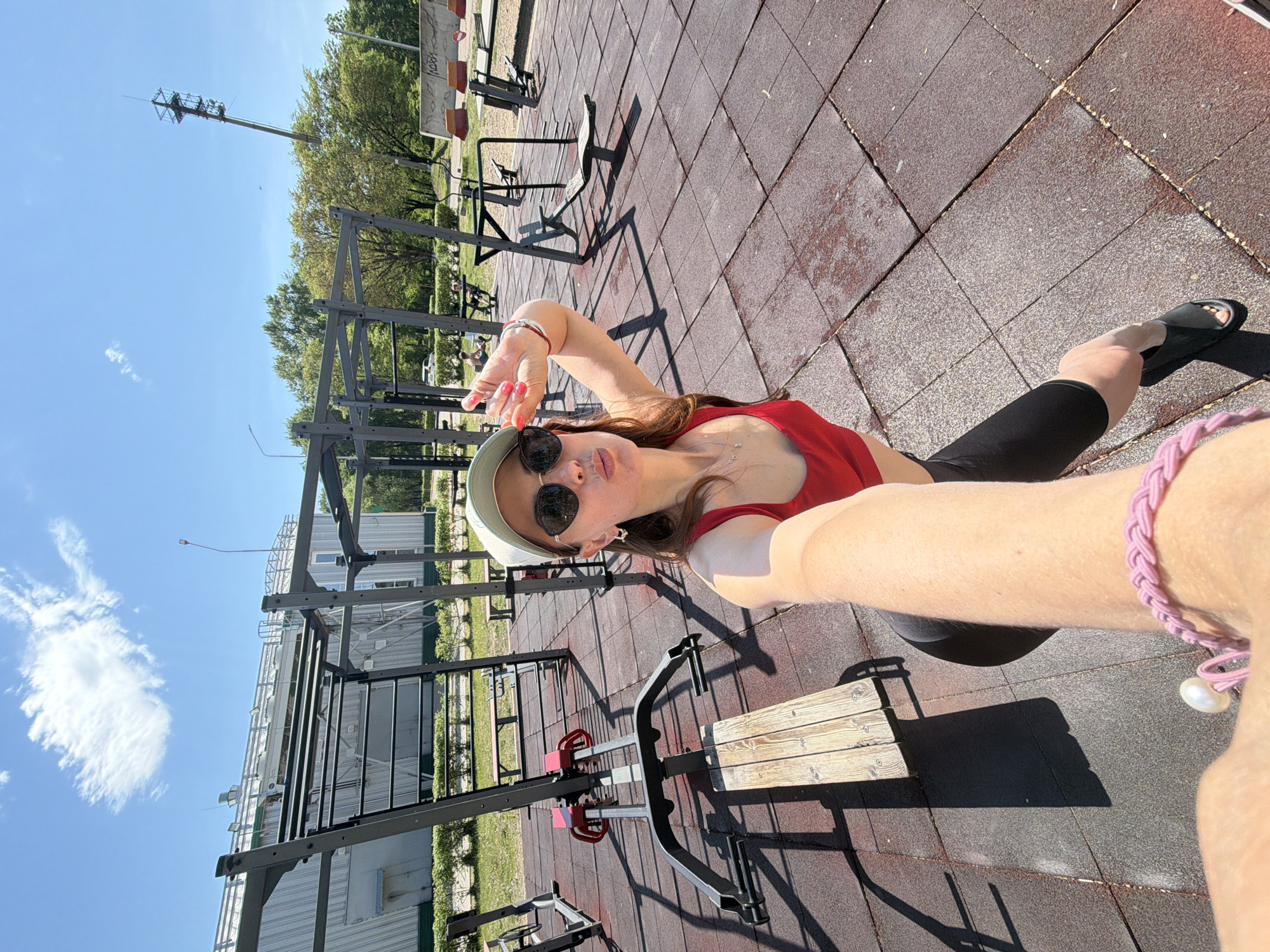 The image shows a young woman in an outdoor gym area in Birobidzhan, Russia. She is wearing a red tank top, black leggings, sunglasses, and a white cap. She is taking a selfie, holding her arm out towards the camera. The gym area has rubber flooring and several pieces of metal exercise equipment. In the background, there are trees, a building, and a communication tower. The sky is blue with some clouds.