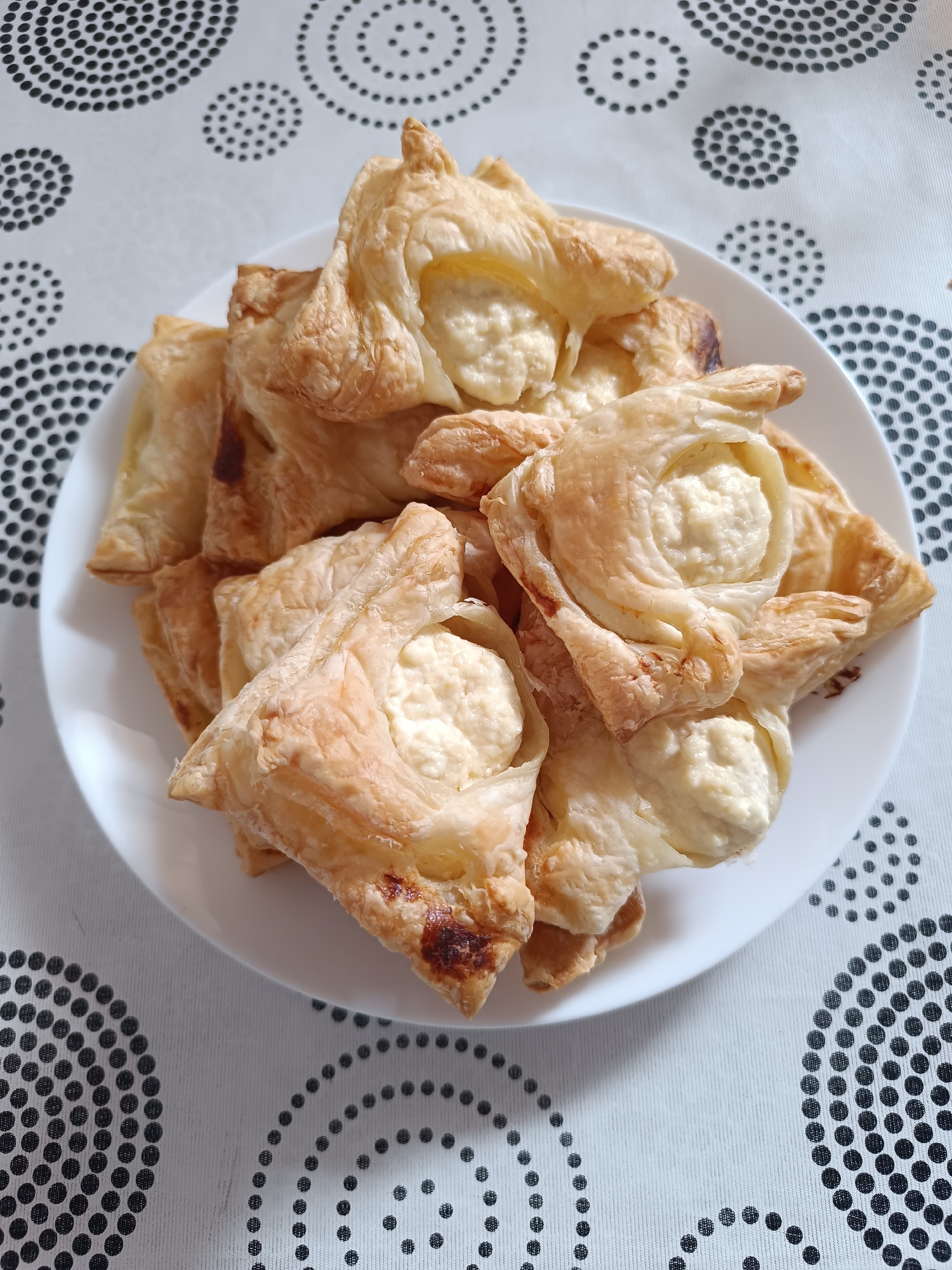 A close-up, overhead shot showcases a white plate filled with golden-brown, flaky pastries. These pastries appear to be filled with a creamy, white substance, likely cheese or a sweet filling. The pastries are arranged in a slightly overlapping manner, with some folded into a quatrefoil shape, exposing the filling in the center, while others are more simply folded into squares. The pastry dough exhibits a puffed and layered texture, indicating it might be puff pastry or a similar laminated dough. Some areas of the pastry show a slightly darker, caramelized brown, suggesting they are well-baked.
The plate rests on a white tablecloth patterned with concentric circles of black dots, creating a visually interesting background that contrasts with the uniform white of the plate and the varied textures of the pastries. The lighting suggests it is daytime, possibly indoors, as there are no strong shadows indicating direct sunlight. The overall impression is one of a home-prepared treat, ready to be served or enjoyed. There is no visible text in the image.
