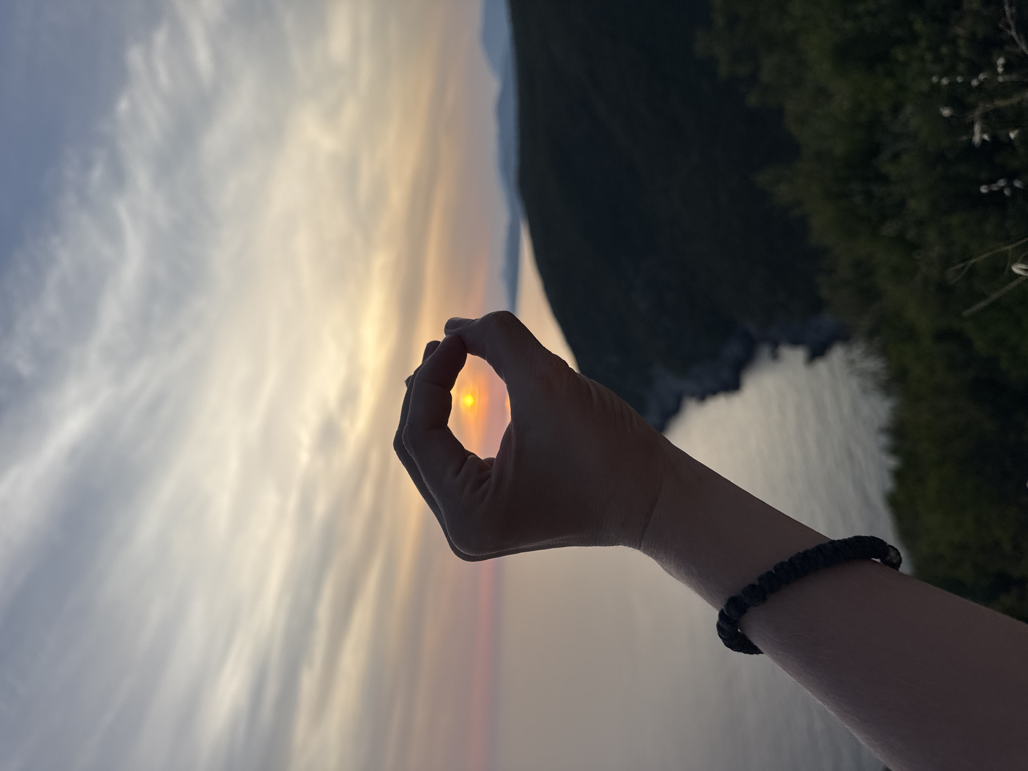 A close-up of a person's hand forms an "O" shape, framing the setting sun over the Adriatic Sea. The sun is a bright orange orb visible through the fingers. The hand is positioned in the foreground, silhouetted against the sea and sky. The person is wearing a dark, braided bracelet on their wrist. In the background, a hilly coastline with dark vegetation is visible, with faint mountain ranges in the distance. The sky is filled with wispy clouds in shades of gray, orange, and yellow, indicating a sunset. The scene is located near Zagora, Montenegro, along the coastline.