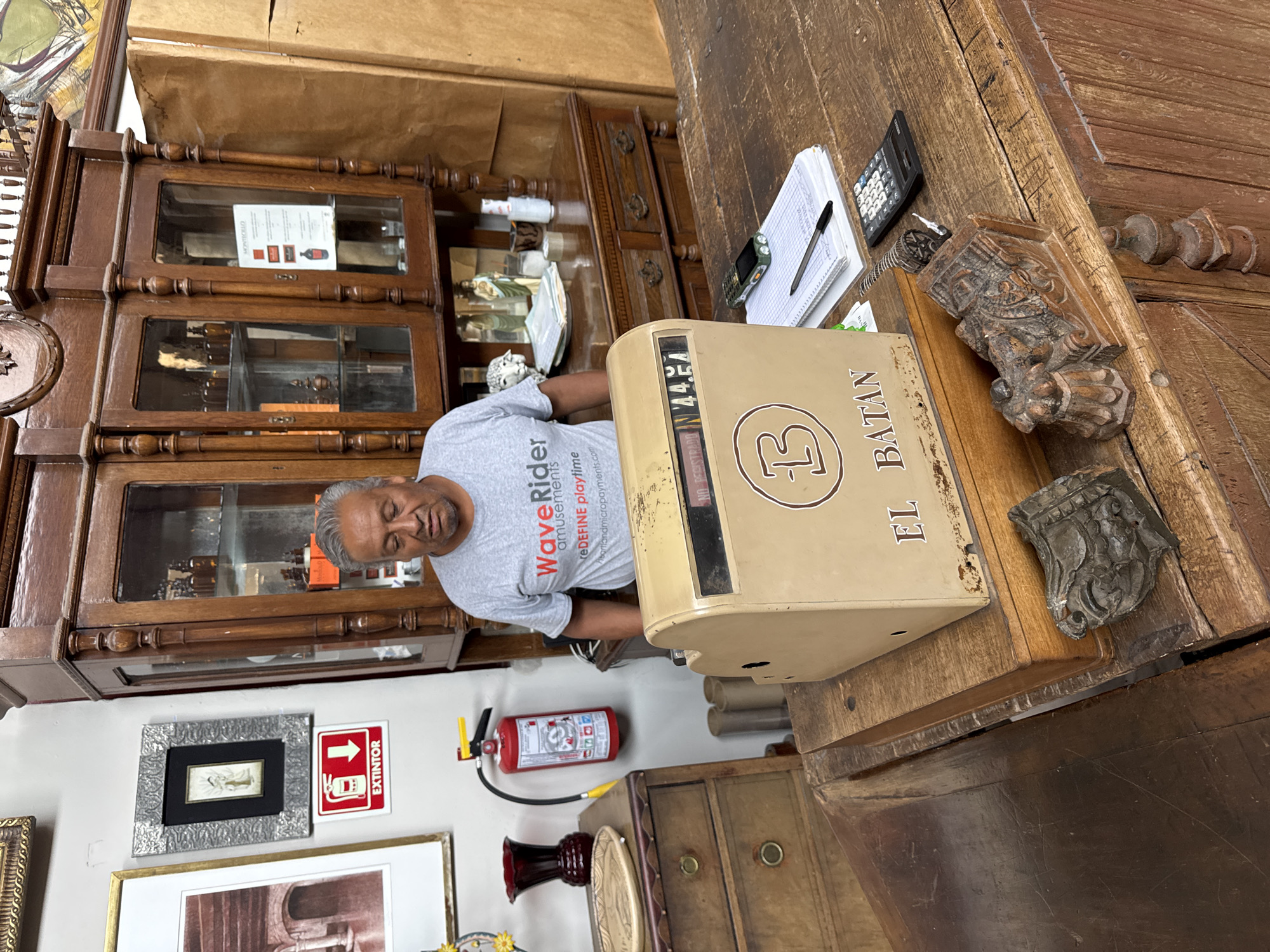 A man is standing inside a shop. He is wearing a grey t-shirt with "Wave Rider amusements" written on it. In front of him on a wooden counter is an antique cash register with "EL BATAN" printed on its side. Next to the cash register is a calculator and notepad. The counter is made of dark, carved wood. Behind the man is a wooden cabinet with glass doors containing various objects, including bottles and decorative items. On the wall next to the cabinet is a framed picture, a sign with "EXTINTOR" written on it, and a fire extinguisher. This is likely in San Miguel de Allende, Mexico, given that EL BATAN is a popular Mexican restaurant chain.