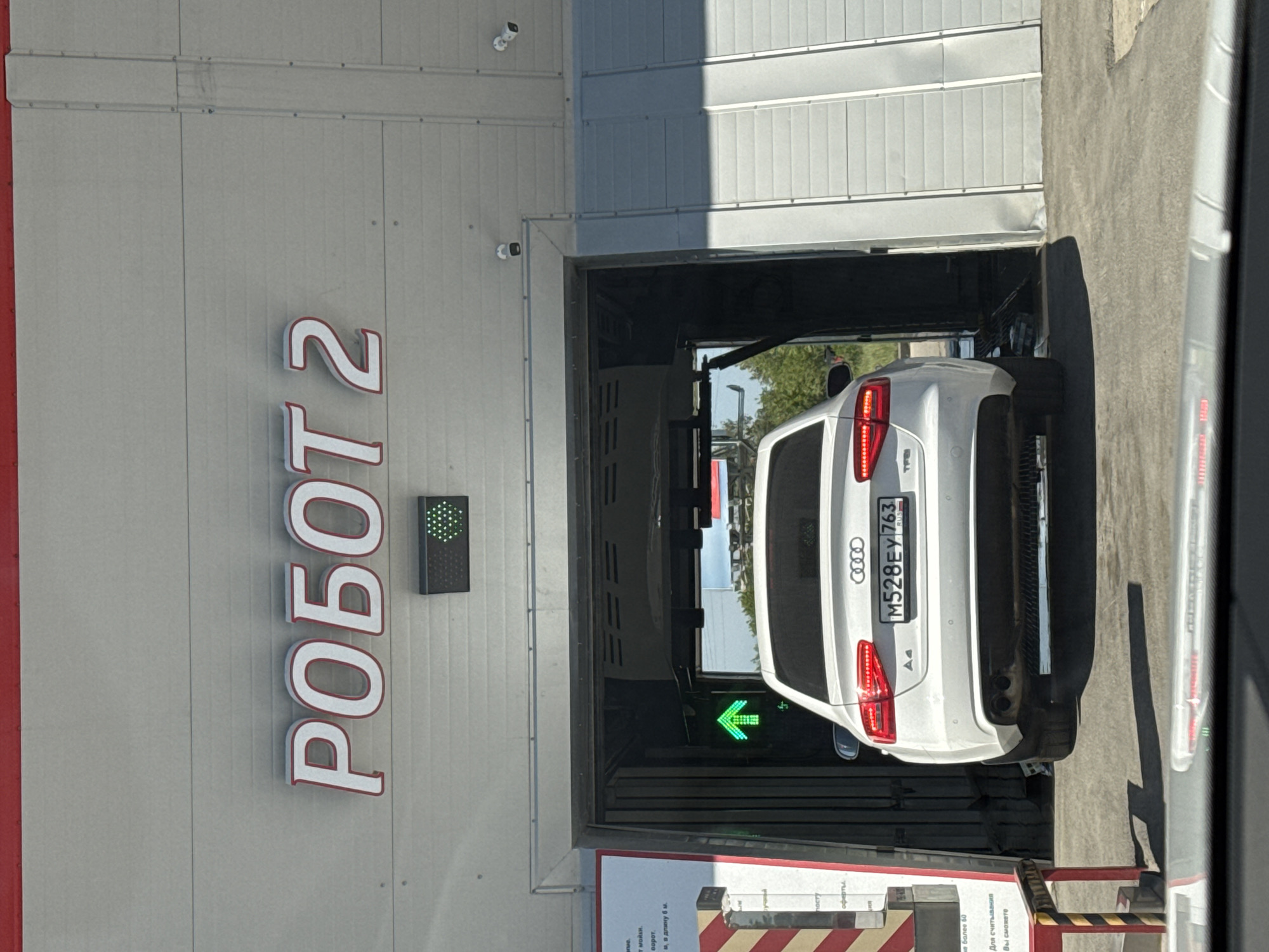 Here's a detailed description of the image:
**General Overview:**
The image shows a white Audi A4 car entering a car wash labeled "РОБОТ 2." The scene is likely in an urban area, judging by the commercial setting. The photo appears to be taken from inside another vehicle, looking out through a window.
**Specific Elements:**
* **Car Wash:** The car wash entrance is visible. The name "РОБОТ 2" is displayed vertically in large, red letters with a white outline on the side of the building.
* **Car:** A white Audi A4 is entering the car wash. The license plate number is M528EY 763. There's a green arrow signal inside the car wash, indicating the direction to proceed.
* **Building Exterior:** The building appears to be made of grey corrugated panels. There are surveillance cameras mounted near the entrance.
* **Environment:** The setting appears to be a bright, sunny day.
* **Other Signage:** Red and white striped safety barricades.
* **Location:** City of Unknown, Unknown