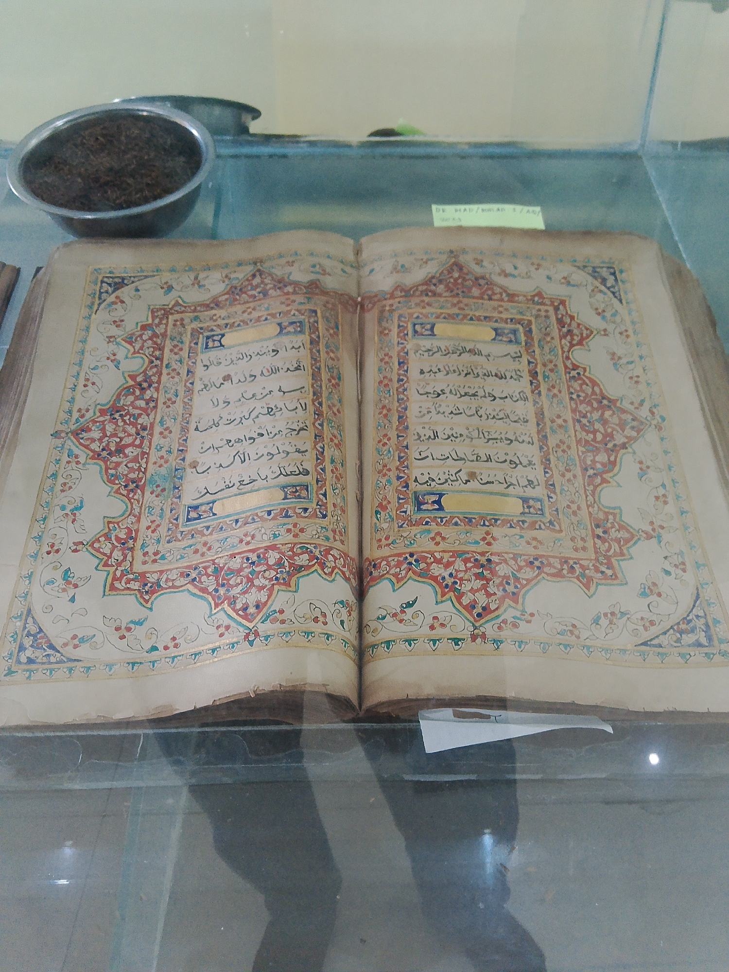 The image depicts an open, antique book with ornate decorations on both pages. The book is lying on a flat surface, likely a glass display case. In the upper left corner, there is a metal bowl filled with dark, granular material, possibly tea leaves or spices. Behind the bowl, another dark object is visible. A small white piece of paper with some writing is attached to the glass in front of the book. The book's pages are aged and slightly yellowed. The calligraphy within the book is elaborate and appears to be in Arabic. The background is a light-colored wall.
Based on the text visible on the white paper in the image, "DR. KH. ABDUL HALIM / AM...", and cross-referencing with information about prominent Islamic scholars and historical figures from Demak, Indonesia, it is highly probable that this display is located at the "Masjid Agung Demak" (Great Mosque of Demak) or within its museum/cultural heritage complex. Masjid Agung Demak is a significant historical mosque in Demak, Central Java, Indonesia, known for its role in the spread of Islam in Java and its historical artifacts. The presence of an antique book, possibly a religious text, aligns with the museum-like exhibits often found in such historical religious sites.