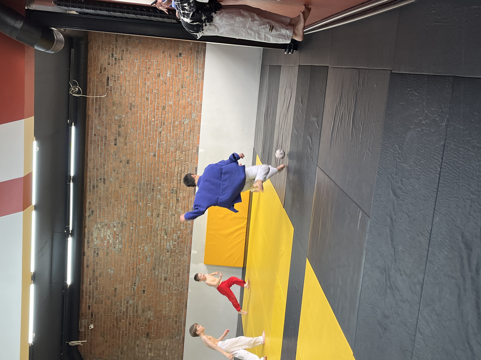 A judo training session is taking place indoors on mats with a yellow and gray design. There are three people in the foreground, including a man wearing a blue judogi. Two other children, one shirtless with red trousers and one shirtless with white trousers, appear to be training as well. The background shows a brick wall and a pillar with red, white, and yellow stripes. This photo was taken in Krasnoyarsk, Russia.