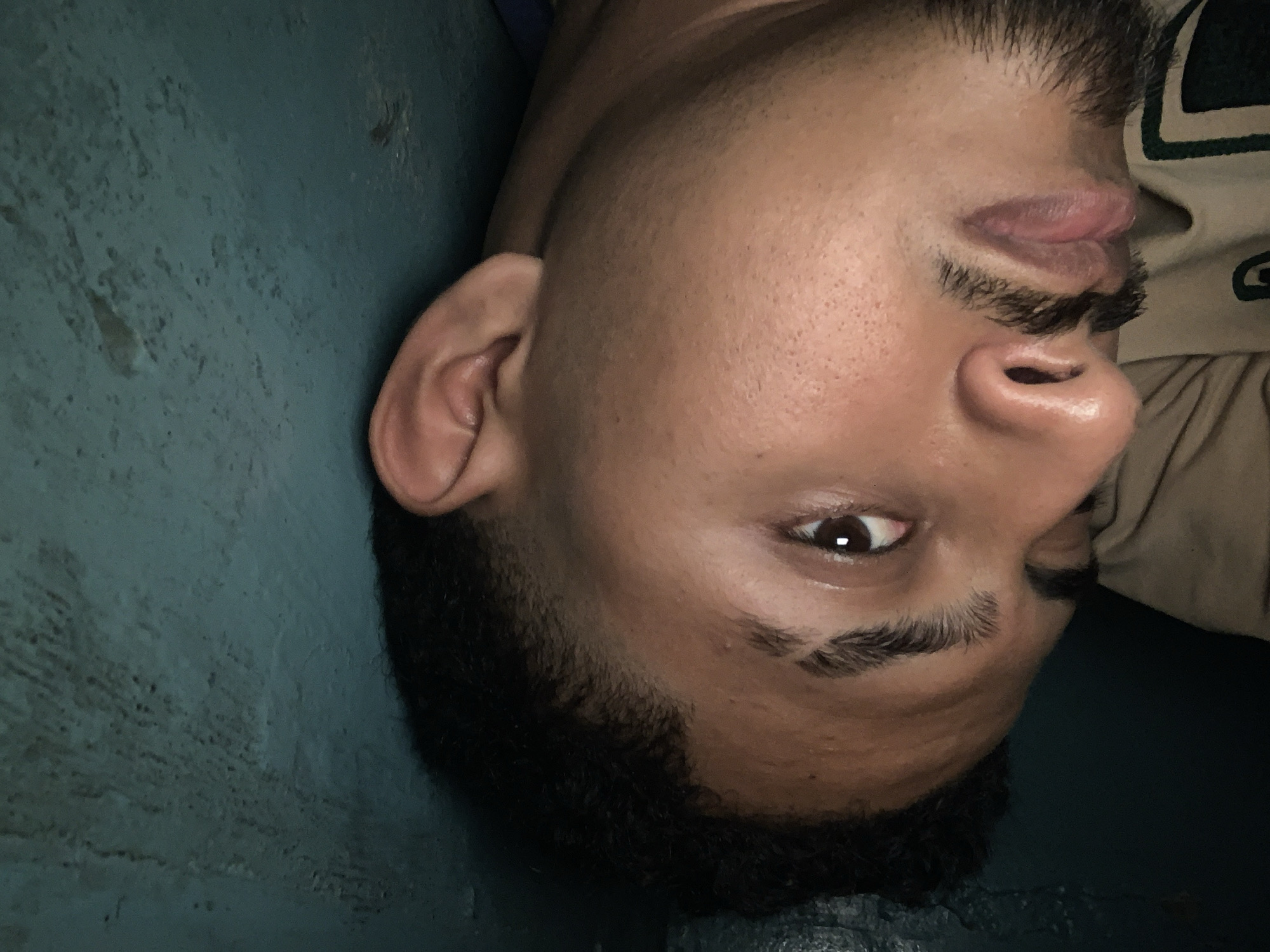 A close-up portrait displays a young man's face, rotated approximately 90 degrees clockwise, giving an inverted perspective. He has dark skin, short dark hair, and a trimmed dark beard and mustache. His right eye is open, gazing directly towards the camera, and his lips are slightly parted. He is wearing a light-colored shirt, possibly beige or cream, with a dark green or black graphic on it that includes a partial white letter, appearing to be a stylized "G".
The background consists of a textured wall, colored in a muted greenish-blue or teal, showing signs of wear or an uneven surface. The lighting is somewhat dim, suggesting an indoor setting or low-light conditions. The man's expression is neutral, indicating he is posing for the photograph. The scene focuses solely on the man's face and the immediate wall behind him. The location context provided is Santa Ana, Venezuela.