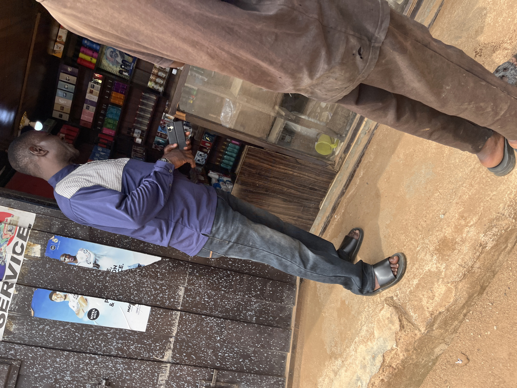 The image captures a street scene in Jos, Nigeria, likely in a commercial area. In the foreground, two men are partially visible. The man on the left is wearing a blue collared shirt with a checkered white and gray pattern on the upper chest and shoulders, dark wash jeans, and black slide sandals. He is holding a black smartphone in his right hand and appears to be looking at it or the shop in front of him. The man on the right is mostly obscured, wearing a brown t-shirt and brown pants, and black slide sandals.
The background features a small shop with shelves filled with various products, possibly toiletries or cosmetics, identifiable by colorful packaging and bottles. A glass display case is in front of the shelves. To the left of the shop, a weathered brown metal door is visible, adorned with two posters. One poster shows two male athletes in white sports attire, and the other appears to be an advertisement for NIVEA MEN products. The ground is a reddish-brown, dusty earth, suggesting a dry environment. The overall impression is of an everyday street vendor or small shop in an urban setting.