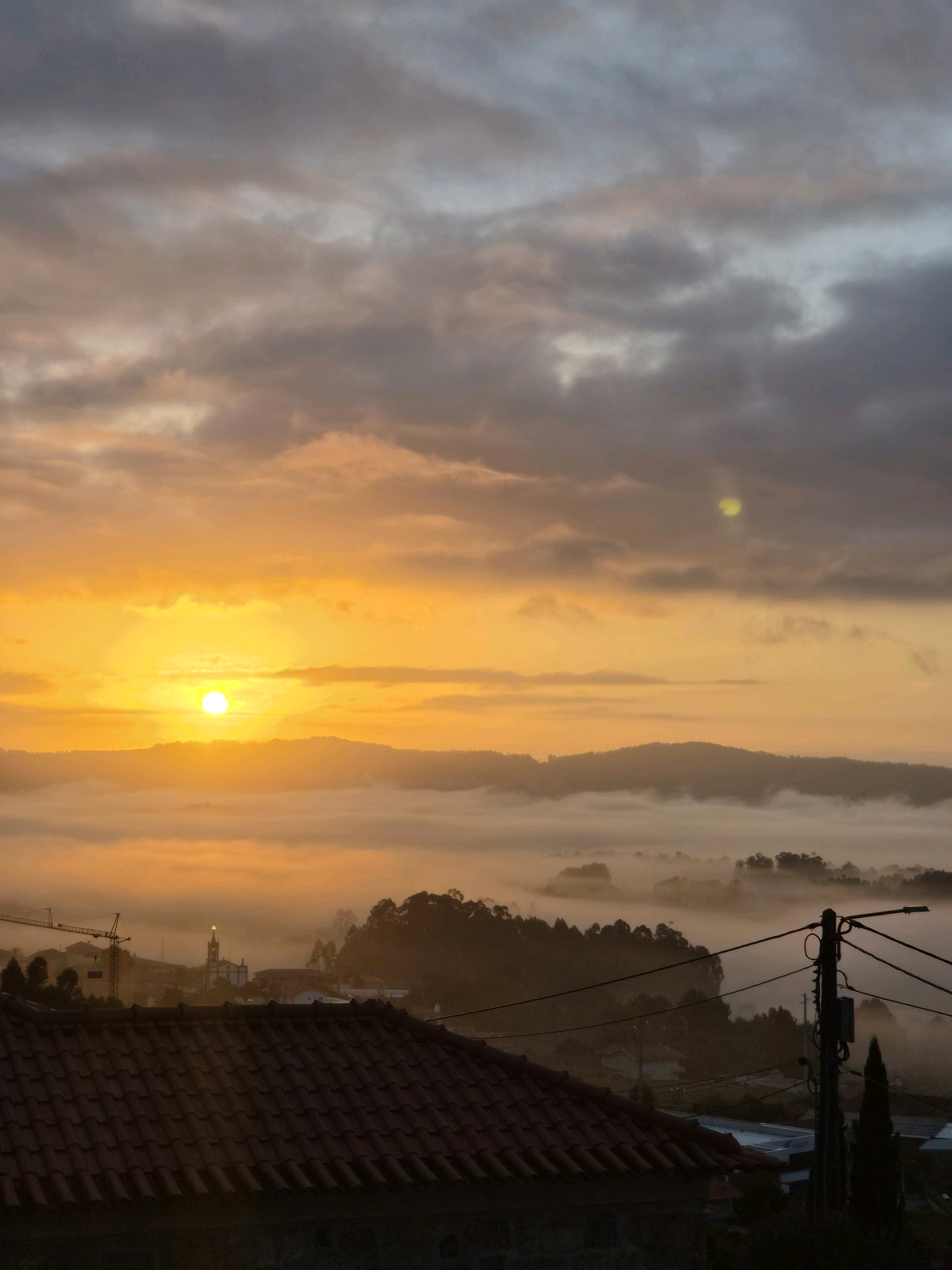 The image depicts a sunrise over the city of Barcelos, Portugal. The sun, a bright orange orb, is rising over a line of misty hills. The sky is filled with clouds in various shades of orange, yellow, and gray. In the foreground, there is a tiled roof of a building. Beyond the roof, a town is visible, with houses and a church steeple. A construction crane can be seen on the left side of the image. Power lines and a utility pole are also present in the foreground, to the right. The overall scene is tranquil and atmospheric, with the mist and the sunrise creating a soft, ethereal light.