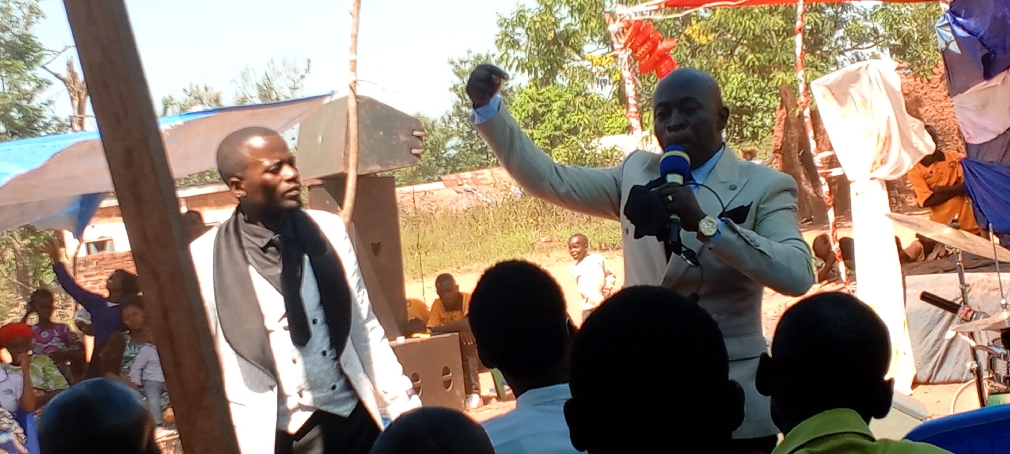 The image depicts a gathering, possibly a church service or event, in an outdoor setting. It is daytime with bright sunlight. In the foreground, individuals are looking at the camera.
A man in a white suit holds a microphone. Beside him stands another man in a white vest and dark clothing. Both appear to be the focal points of attention.
In the background, a crowd is seated. The setting suggests a makeshift outdoor venue, with tent-like structures erected using wooden poles and draped fabric. Musical instruments are visible on the right, indicating live music.
The location is Kalemie, Democratic Republic of the Congo, based on the visual characteristics of the scene.