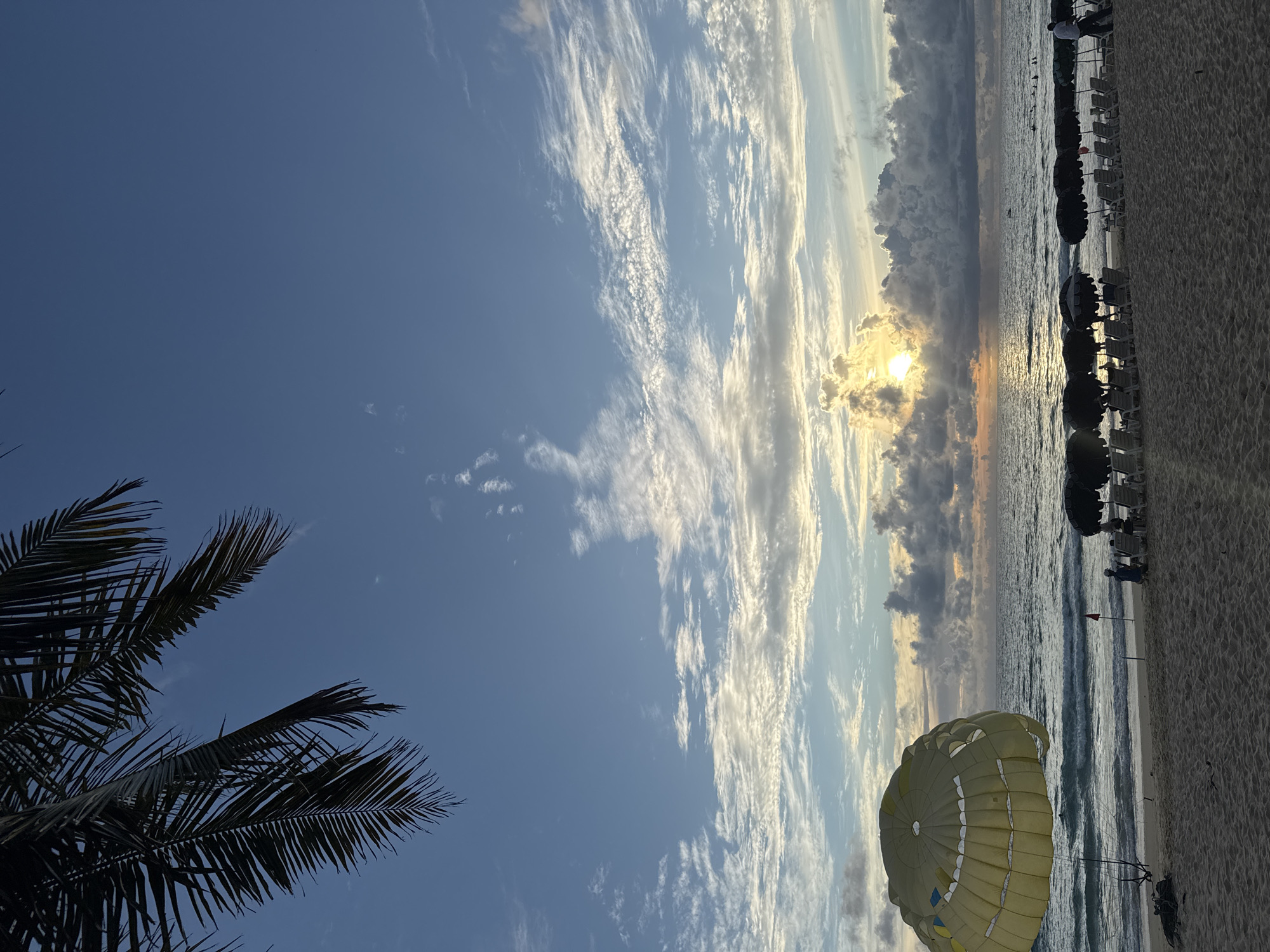 This image captures a beach scene at what appears to be sunset or sunrise, with the sun peeking through dramatic clouds over the ocean. In the foreground, a large, yellow parachute is laid out on the sandy beach, suggesting an activity like parasailing is either about to happen or has just concluded. To the left, the dark silhouette of palm fronds frames the sky.
The beach itself is lined with rows of empty lounge chairs and umbrellas, indicating a resort or a popular tourist spot. The ocean has gentle waves, and the sky is a mix of deep blue with streaks of white and gray clouds, illuminated by the sun's warm glow. The overall mood is serene and tranquil, with a hint of adventure due to the parachute. No text is visible in the image. The setting is likely a tropical beach, and given the context provided, it could be Karon, Thailand.