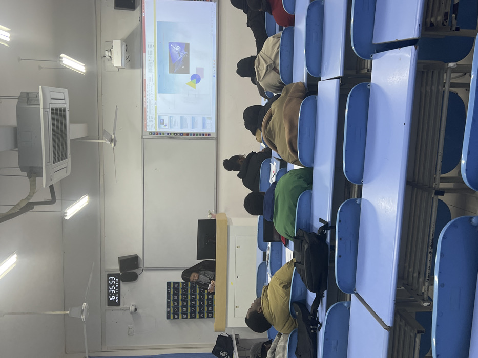 The scene depicts the interior of a classroom or lecture hall in Jiangjin, China, observed from a mid-to-rear perspective. At the front of the room, a female individual with dark hair, wearing a grey top and dark jacket, stands behind a light-colored podium. She is looking down at a small electronic device held in her hands. A black computer monitor is positioned on the podium. Behind her, a large white board spans the wall. To the right, a presentation is projected onto a screen, displaying what appears to be a PowerPoint slide featuring an image (possibly of machinery or a satellite) alongside a yellow triangle, a blue partial circle, and a red rectangle. A projector is ceiling-mounted, casting the image.
On the left wall, a digital clock displays "16:35:53" and a date (202*x*年1月19日). Below the clock, a white security camera is mounted, and below that, a grid-like display with numbers arranged in columns (e.g., 01-06, 07-12). Two black wall-mounted speakers are positioned above the whiteboard.
The room is illuminated by multiple rectangular fluorescent light fixtures and features two three-bladed ceiling fans. A large, rectangular ceiling-mounted air conditioning unit is also visible, with ducts running across the ceiling.
In the foreground and midground, approximately ten individuals, likely students, are seated in light blue desks with attached chairs. Most are viewed from behind. Several students are wearing winter jackets in various colors, including mustard yellow, green, and black. One male individual in a mustard yellow jacket in the second row is leaning back, appearing to be asleep or resting with his eyes closed. A black laptop is open on a desk belonging to a student in a green jacket in the second row. A black backpack is placed on an empty desk in the front row. The students' actions vary, with some looking towards the front and others focused downwards on their desks.