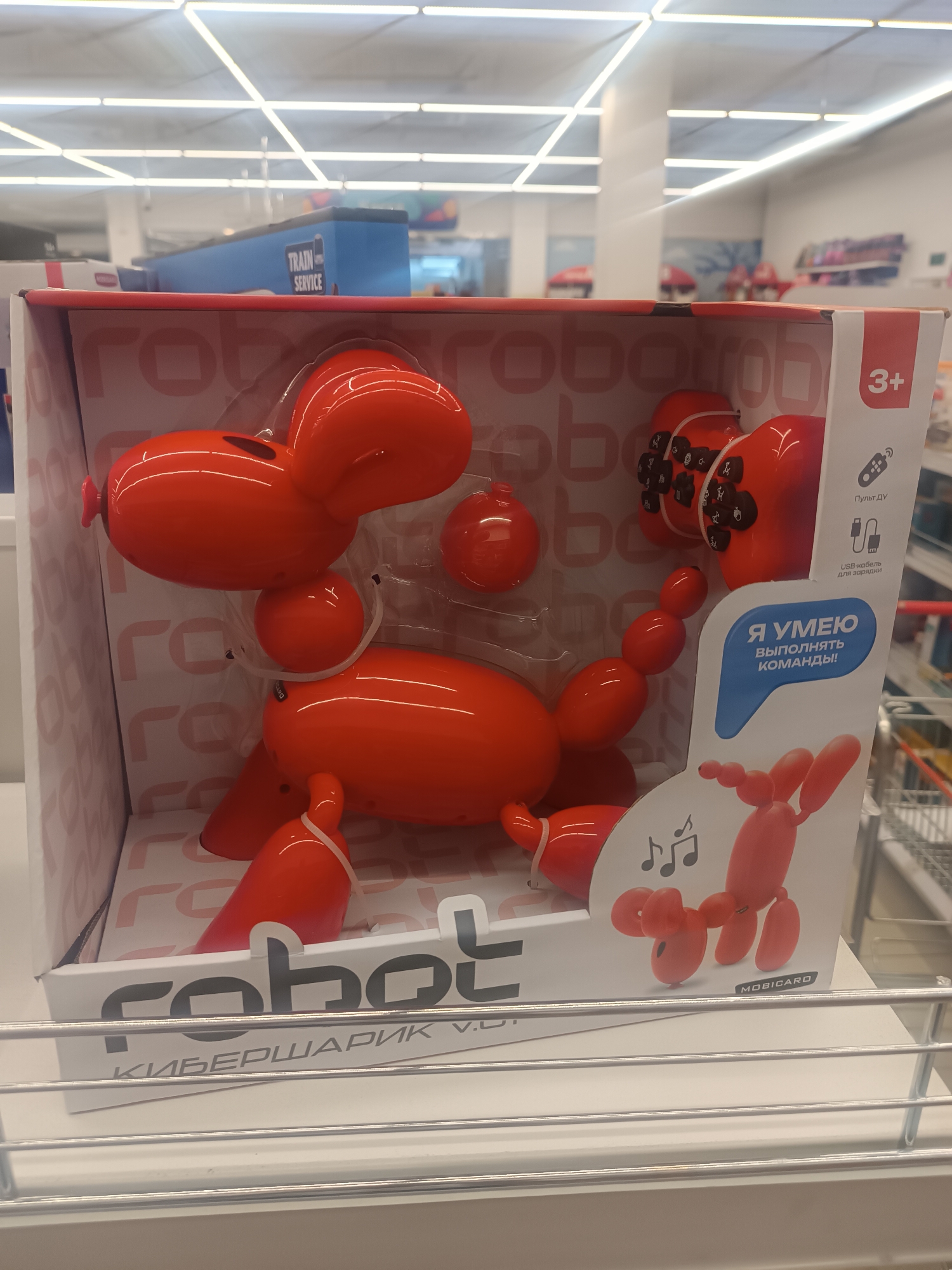 An orange robotic toy dog, resembling a balloon animal, is displayed in its original packaging on a retail shelf. The packaging is primarily white with red accents and features text in Russian. The toy dog has segmented body parts and appears to be made of plastic. The box also shows a remote control icon and a USB charging cable icon. On the right side of the packaging, text in Russian states "Я УМЕЮ ВЫПОЛНЯТЬ КОМАНДЫ!" which translates to "I CAN FOLLOW COMMANDS!". Below this, a smaller illustration depicts a similar toy dog. The background of the image shows a retail store environment with fluorescent lighting and shelves stocked with various products. Specifically, a blue sign with "TRAIN SERVICE" is visible in the upper left portion of the image, suggesting this store might be located in a shopping center or a mall in Tula, Russia, potentially near a train station or a store related to transportation services. The overall scene is a typical retail display of a children's toy.