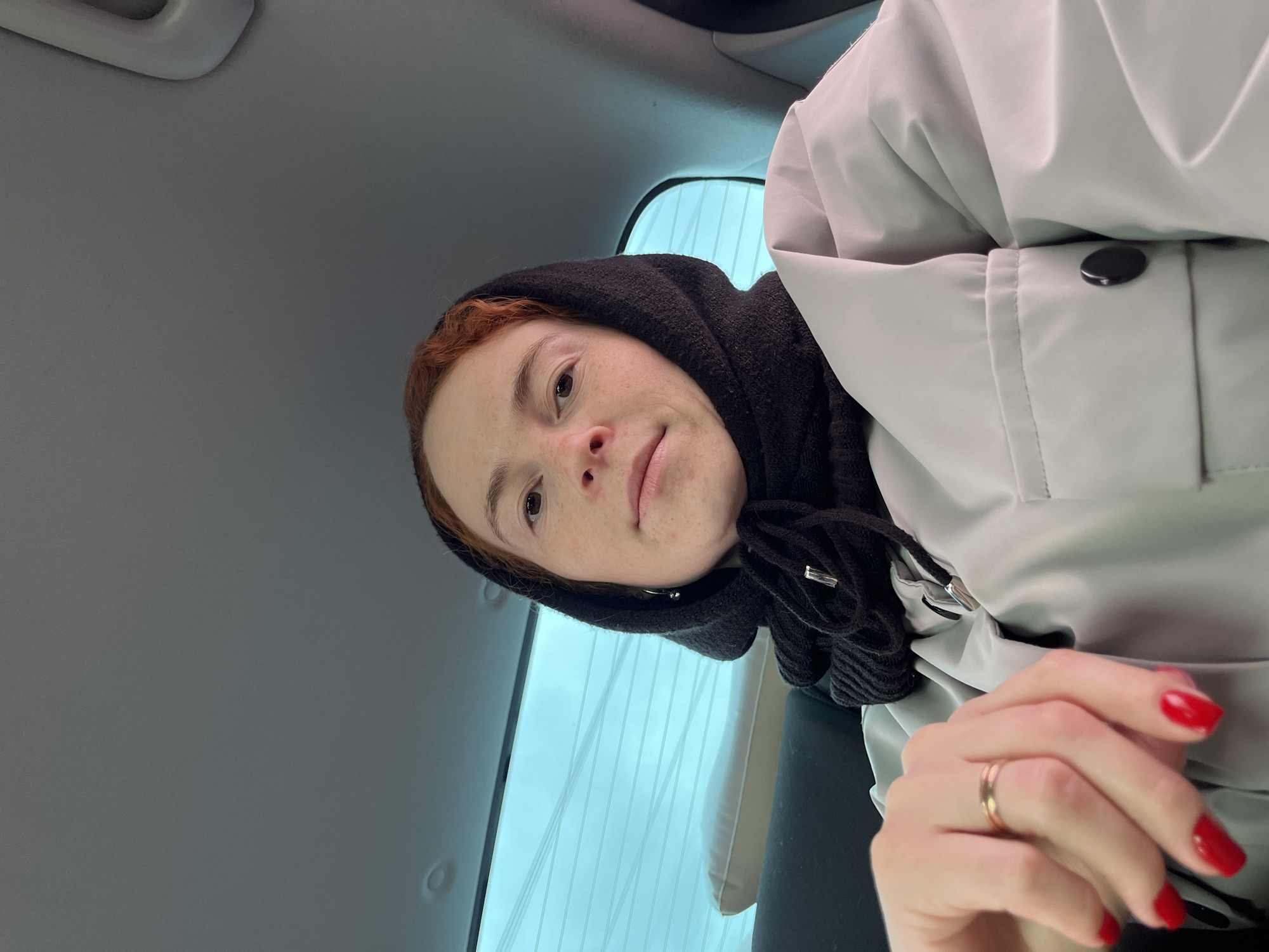 A selfie taken at an oblique angle features a fair-skinned woman with reddish hair, visible freckles, and a slight smile. She is wearing a dark, possibly black, head covering that frames her face. The woman is wearing a light gray or off-white coat or jacket. Her hand, adorned with a gold ring and painted with bright red nail polish, is partially visible in the lower right corner. The background suggests an interior, likely the inside of a vehicle. The rear window behind her is visible, showing what appear to be light-colored vertical lines, possibly blinds or the pattern of defroster lines on the glass. Part of the car’s interior ceiling is visible above her head. The location, Chaikovsky, Russia, cannot be verified from the visual information.