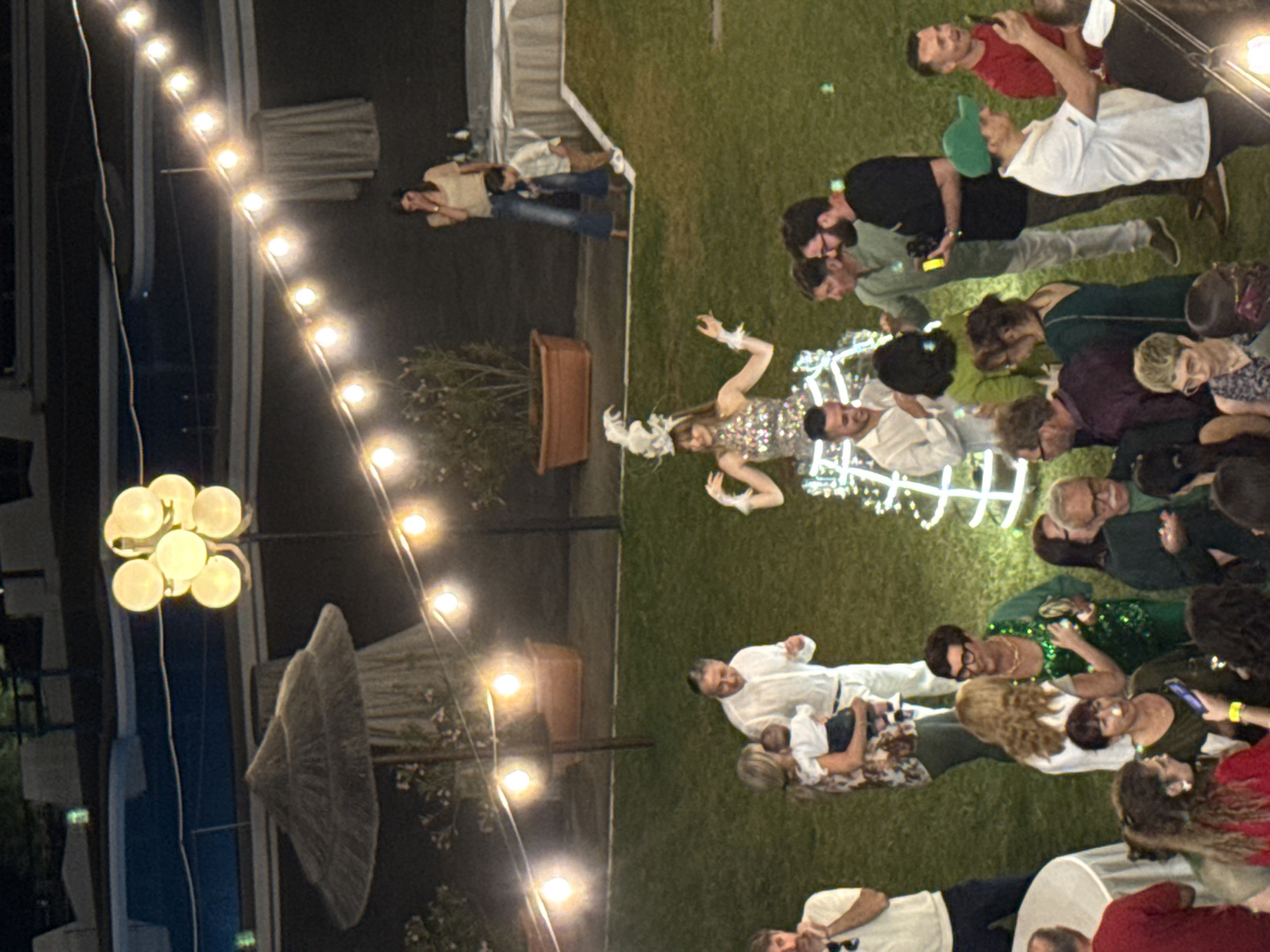 A nighttime outdoor scene in Isola delle Femmine, Italy, features a gathering of people on a lawn. The scene is illuminated by a string of lights and a multi-bulb lamp post. There are around 20 people in view, including a woman in a light-colored, sequined dress with white feathered accessories, who appears to be a focus of the gathering. Other individuals are in casual attire, with some standing and others interacting with each other on the grass. Tables are present, suggesting a party or celebration setting. The environment includes grass, potted plants, and glimpses of buildings or structures in the background.
