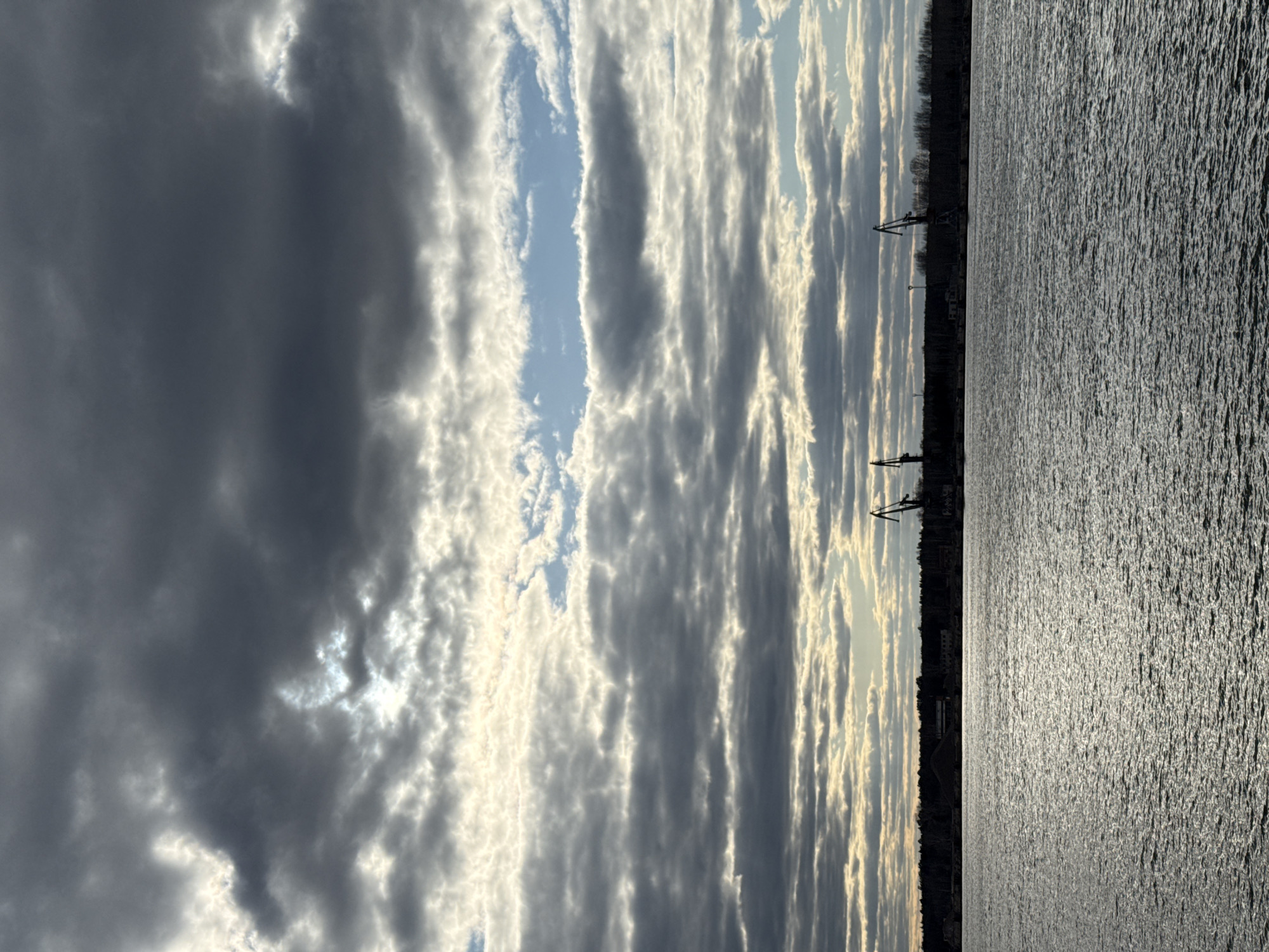 The image shows a body of water, likely the Kama River, adjacent to the city of Chaikovsky, Russia. A line of trees and structures, possibly buildings or industrial facilities, marks the shoreline. Cranes are visible along the edge of the city. The sky is filled with long, striated clouds. Some clouds are dark gray, while others are lighter, allowing sunlight to filter through. The water reflects the light, creating a shimmering effect.