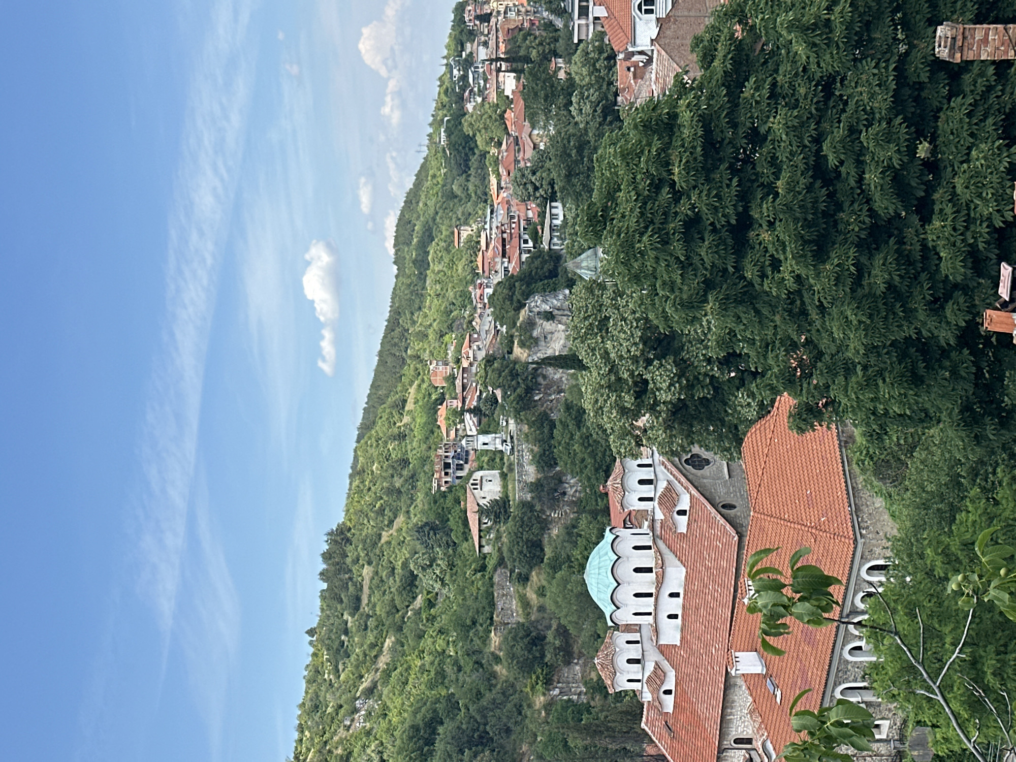 This is an eye-level, medium shot of the city of Asenovgrad, Bulgaria. The photograph showcases a picturesque view of the town nestled against a lush, green hillside under a clear blue sky with scattered white clouds. In the foreground, a distinctive white church with multiple domes, one of which is a striking turquoise, stands prominently with a tiled red roof. This church is likely the St. George's Church, a significant landmark in Asenovgrad.
Behind the church, the city unfolds with a dense collection of buildings featuring terracotta roofs, characteristic of Balkan architecture. Many structures are built into the hillside, some appearing to be residential homes and others possibly part of historical complexes. A white bell tower is visible further up the slope, adding to the architectural tapestry of the city. The surrounding hills are covered in dense green foliage, with trees and shrubs creating a natural backdrop.
The scene appears to be a tranquil, daytime view. No people are visible in the frame, and there are no overt actions or interactions taking place. The overall impression is one of a historic European town set within a beautiful, natural landscape. The presence of the church and the architectural style strongly suggests the location is indeed Asenovgrad, Bulgaria, known for its numerous churches and monasteries.