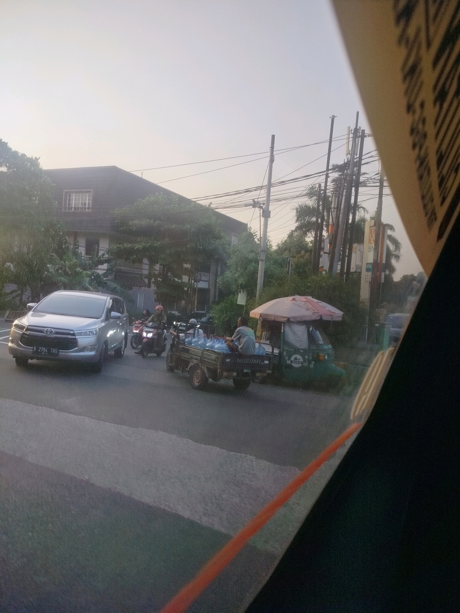 In East Jakarta, Indonesia, on a street with a building in the background, a silver Toyota Innova is parked, its license plate reading "B 2794 TBO". A motorbike with two riders passes by. To the right, a three-wheeled vehicle with an umbrella-covered canopy is stopped, carrying bottles of water in its cargo area. Another person is seated on this vehicle. A utility pole with wires runs across the scene, and trees are visible. The sky is hazy and overcast. A part of a yellow sign with black lettering is visible on the right side of the frame.