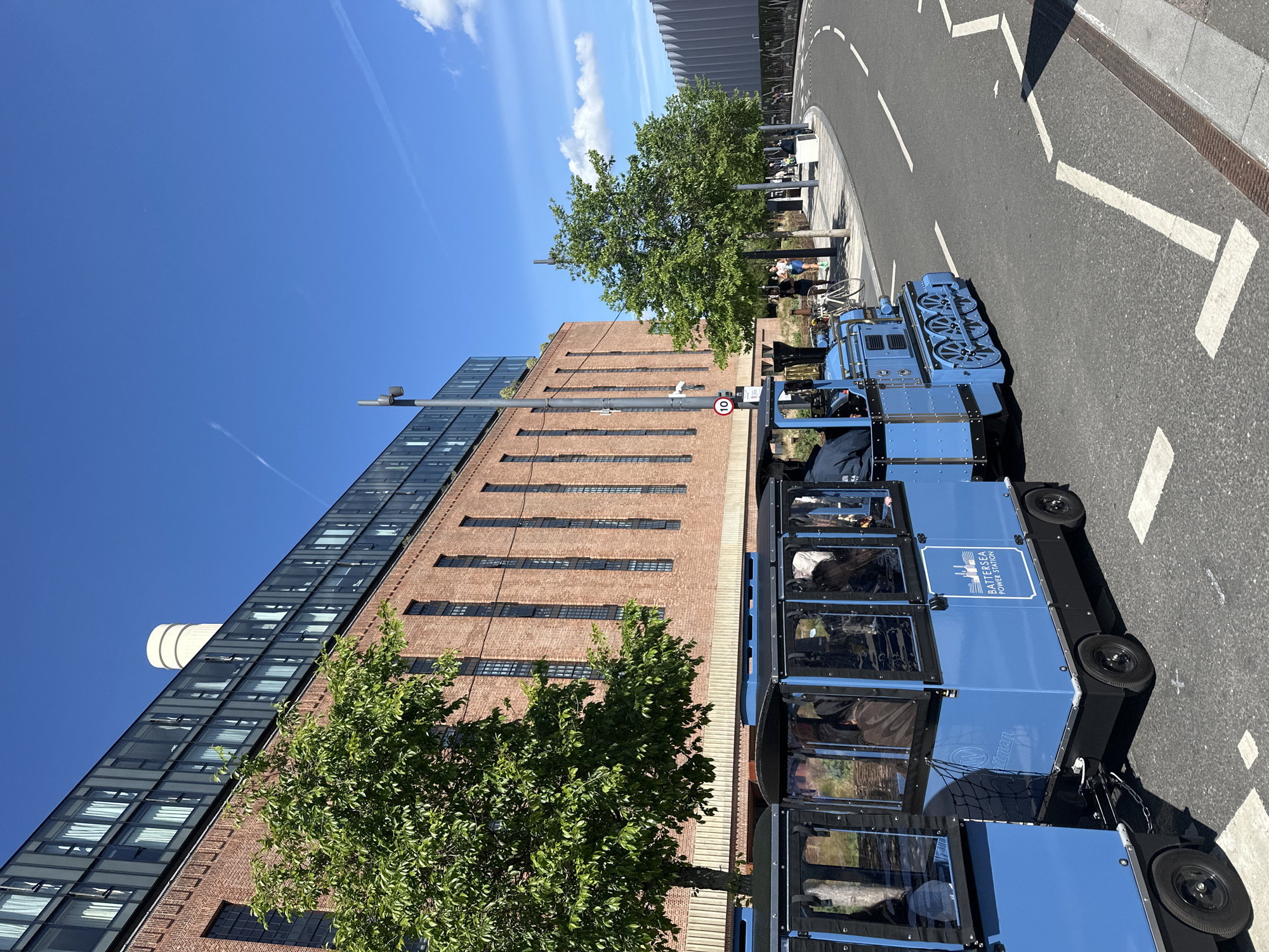 The image depicts a bright, sunny day in London, United Kingdom. A blue, train-like vehicle branded with "BATTERSEA POWER STATION" is parked on a paved street near a building that features a mix of brick and modern glass architecture. The building appears to be multi-storied. A green tree is positioned between the vehicle and the building. Pedestrians can be seen in the background along the curb, and road markings are visible on the street. A sign with the number "10" is present near the tree. The sky is clear blue with a few thin clouds.