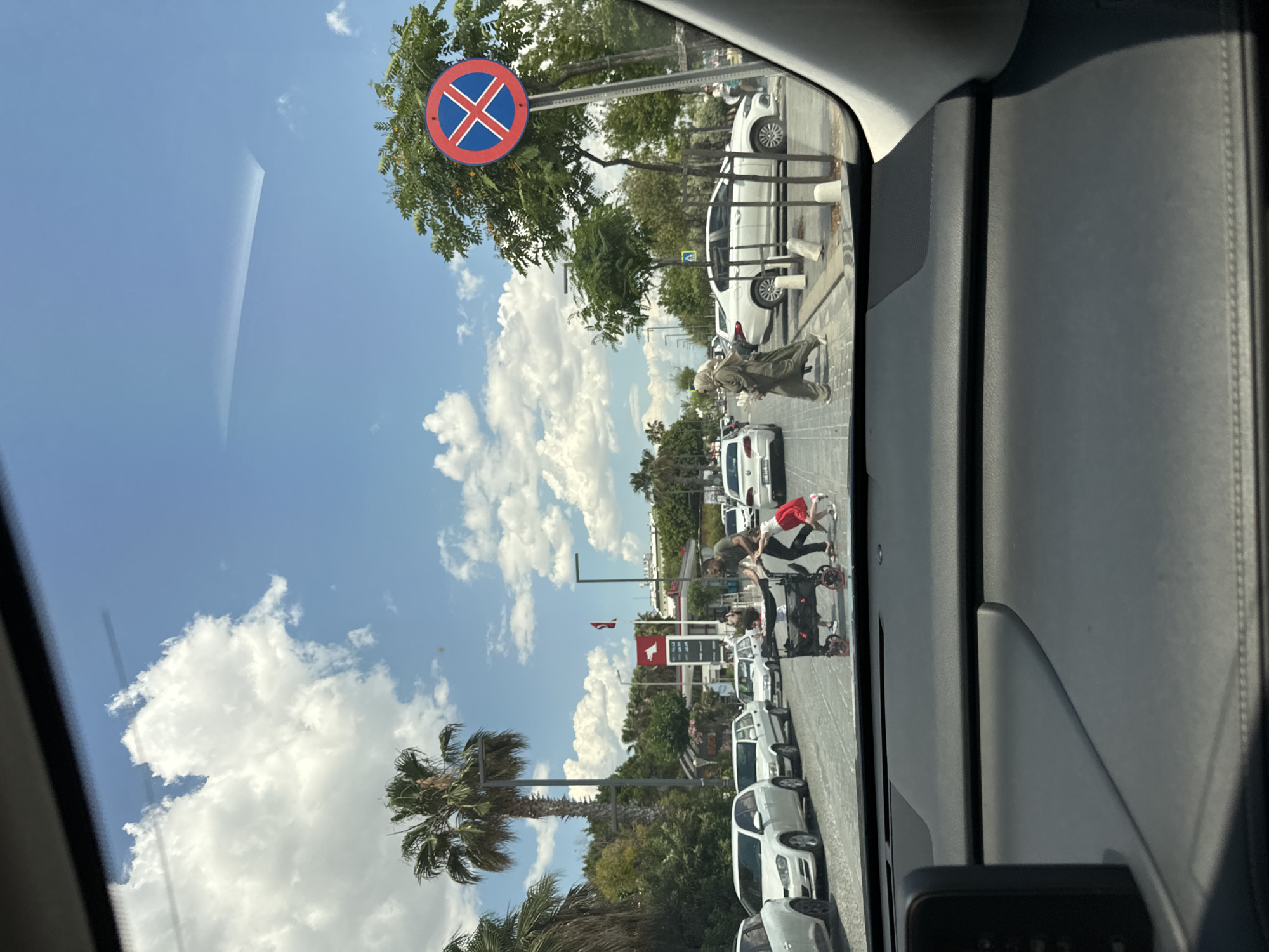 The image, taken from inside a vehicle, shows a street scene in Konyaaltı, Turkey. On the right side of the road are parked white cars, lining a street with palm trees and other green vegetation. A woman in a headscarf is walking toward the street. Further down the street is another person pushing a stroller with a child. A building with a red and white logo is visible. The sky is blue with scattered white clouds. A "No Stopping" road sign is visible on a pole.