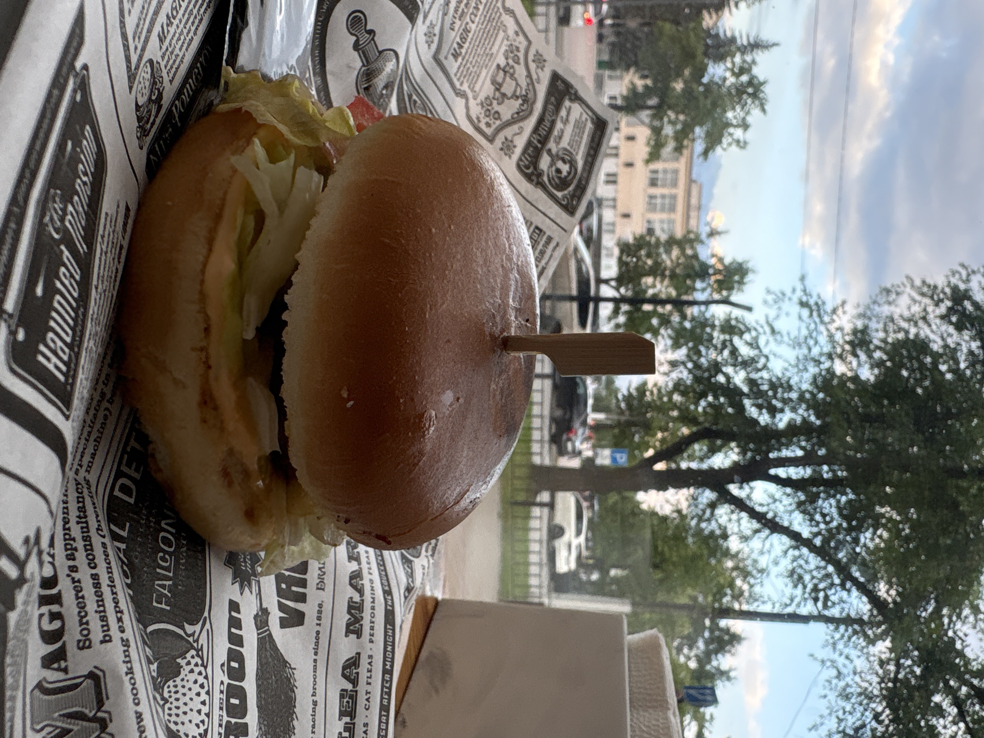 The image displays a close-up of a hamburger presented in a paper wrapper with a newspaper-like print. The hamburger appears to be served with lettuce and possibly tomato, held together by a wooden skewer. The paper wrapper has various text and graphics, including what seems to be a brand name or theme related to "MAGIC" and "Haunted Mansion." In the background, a blurred view through a window reveals trees, buildings, and a cloudy sky, suggesting an outdoor or semi-outdoor setting, possibly a cafe or restaurant. The lighting indicates it is daytime, with soft natural light. There are no people or discernible emotions visible.