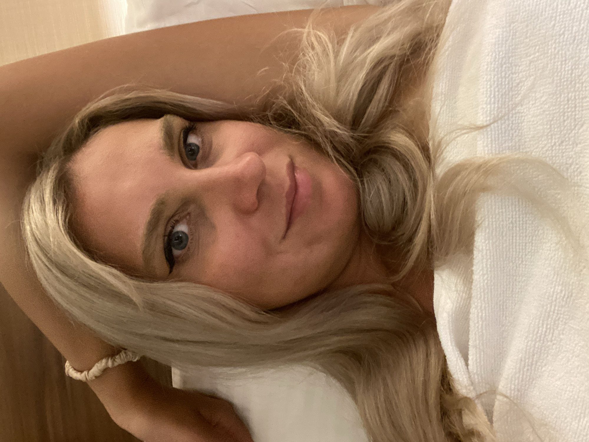 The image is a close-up selfie of a blonde woman lying in bed. She is wearing a white towel or sheet, and her arm is raised above her head. She has blue eyes and is wearing black eyeliner. A beige scrunchie is visible on her wrist. The background is a wooden headboard and a white pillow. There is no information in the image to suggest the location is Aksu, Turkey.
