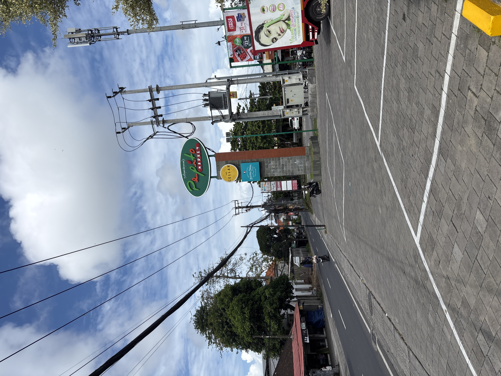 The image shows a street scene in Kuta Selatan, Indonesia. A "Pepito Market" sign is visible on the left. Power lines and poles run along the street. There are storefronts and signs for other businesses. A paved area with white parking space markings is present. A scooter is seen on the road. The sky is partly cloudy.