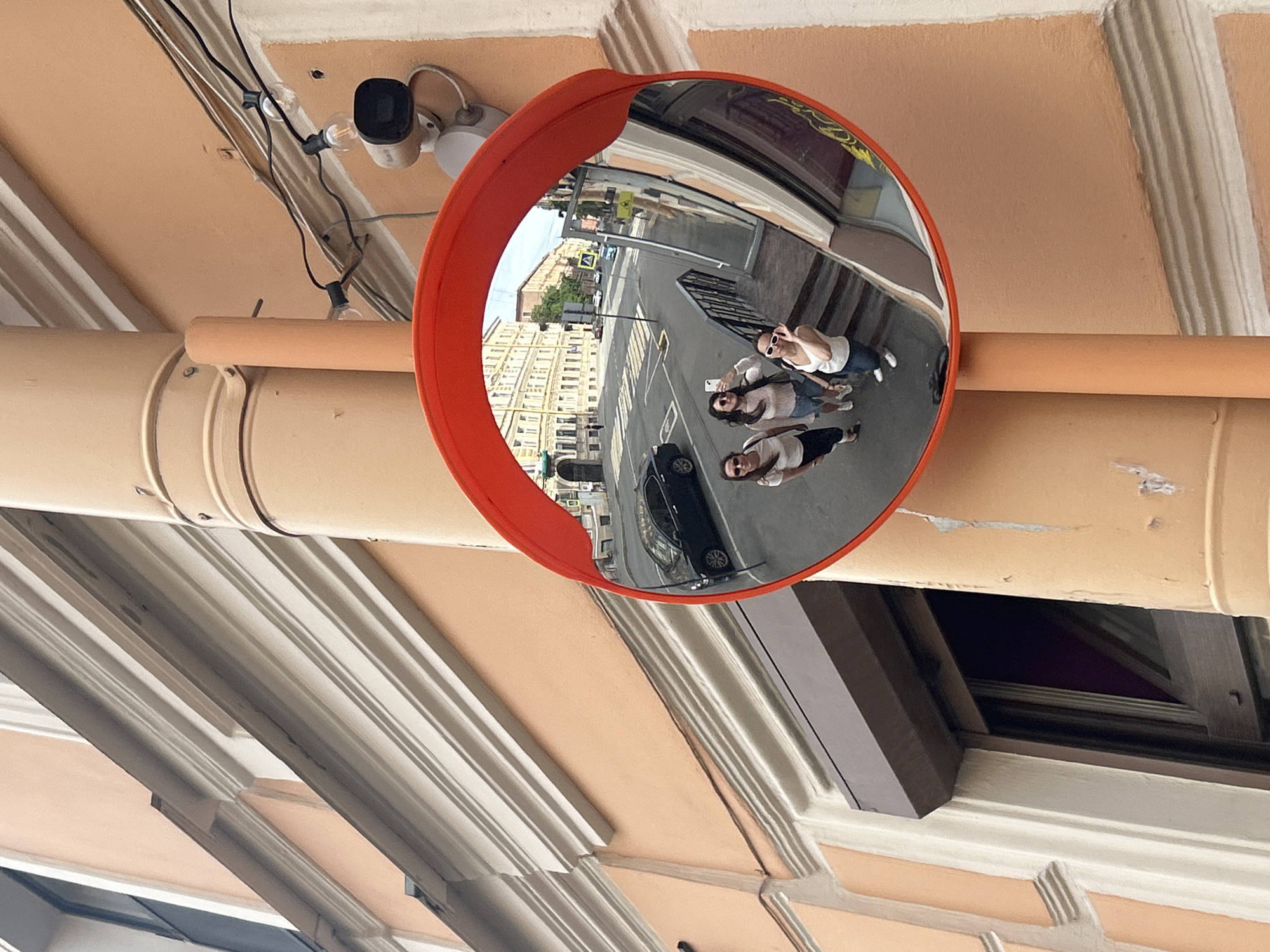 This image depicts a street scene in Saint Petersburg, Russia, featuring a large circular convex mirror with an orange frame, mounted vertically on a beige metal drainpipe. The drainpipe is affixed to a multi-story building, characterized by a peach-colored stucco facade and white decorative architectural moldings. Above the mirror, a white security camera is installed, alongside a string of bare lightbulbs with black wiring. In the lower left foreground, a dark brown roller shutter covers the top of a recessed window or doorway.
Reflected in the convex mirror are three young adult women, positioned to look upwards towards the mirror's perspective, appearing to pose for a photograph. The woman on the left wears a white t-shirt and dark pants with sunglasses pushed onto her head. The central woman, in a light-colored top and light denim shorts, holds a smartphone aloft, seemingly taking a selfie. The woman on the right wears a white sleeveless top and blue jeans, holding white sunglasses. They are standing on steps or a slightly raised pavement.
Behind the women in the reflection, a city street is visible, showing a dark sedan parked, white crosswalk markings, and distant multi-story buildings with classic European architecture under a bright sky.
