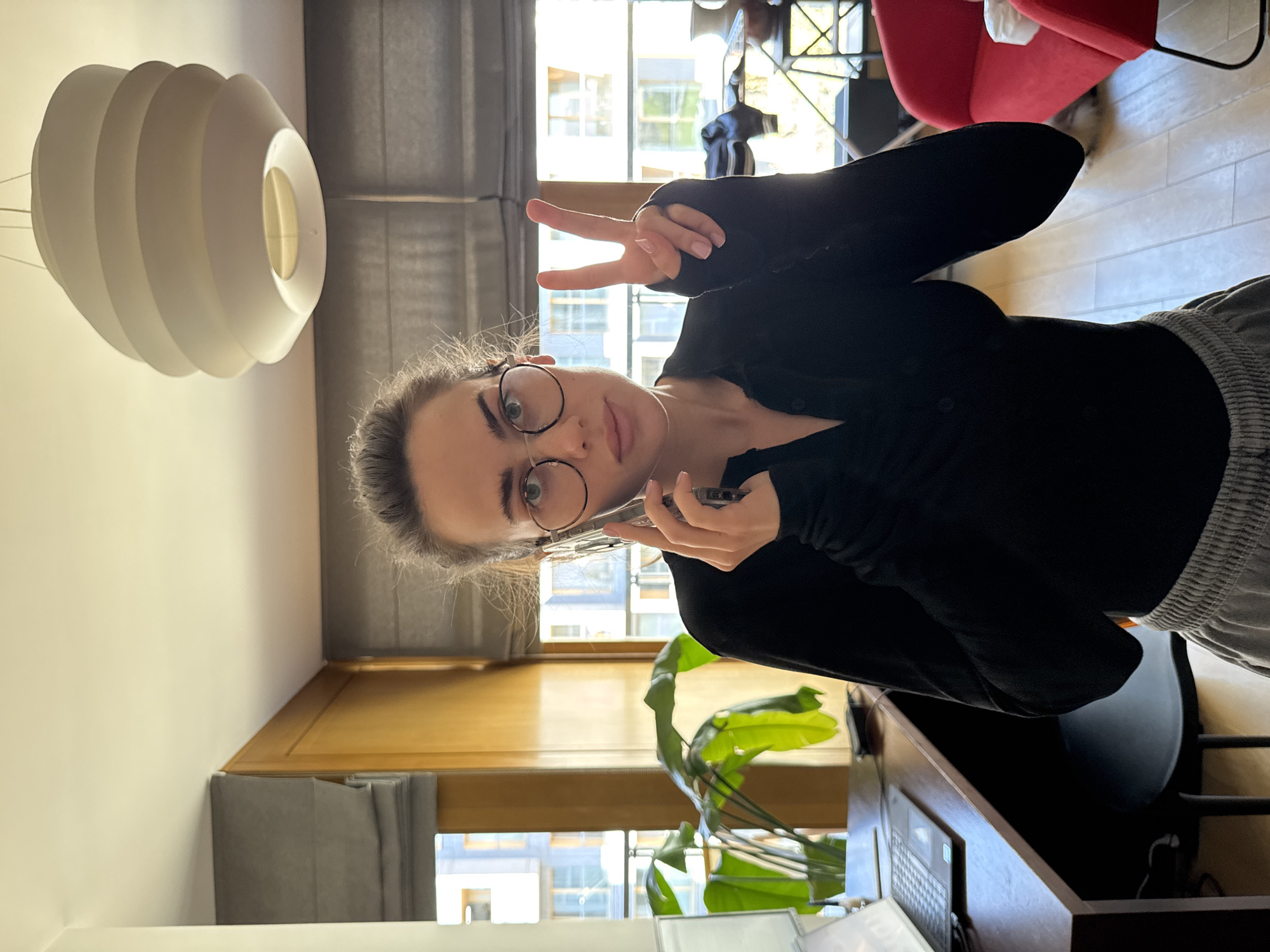 A young woman is depicted indoors, looking directly at the camera with a neutral to slightly smiling expression. She has dark hair pulled back, wears round glasses, a black long-sleeved top, and grey pants. She holds a smartphone to her right ear, suggesting she is on a call, while simultaneously making a peace sign with her left hand.
The setting appears to be a home office or living room, brightly lit by daylight streaming through a large window. Through the window, urban residential buildings are visible, consistent with a city location like Warsaw, Poland. A distinctive white, layered pendant lamp hangs from the ceiling in the foreground. Other visible objects include a large green-leafed plant, a wooden desk with a partial view of a laptop, a red chair, and light-colored floor tiles. The scene conveys a casual, well-lit daytime atmosphere. No discernible text is visible in the image.
