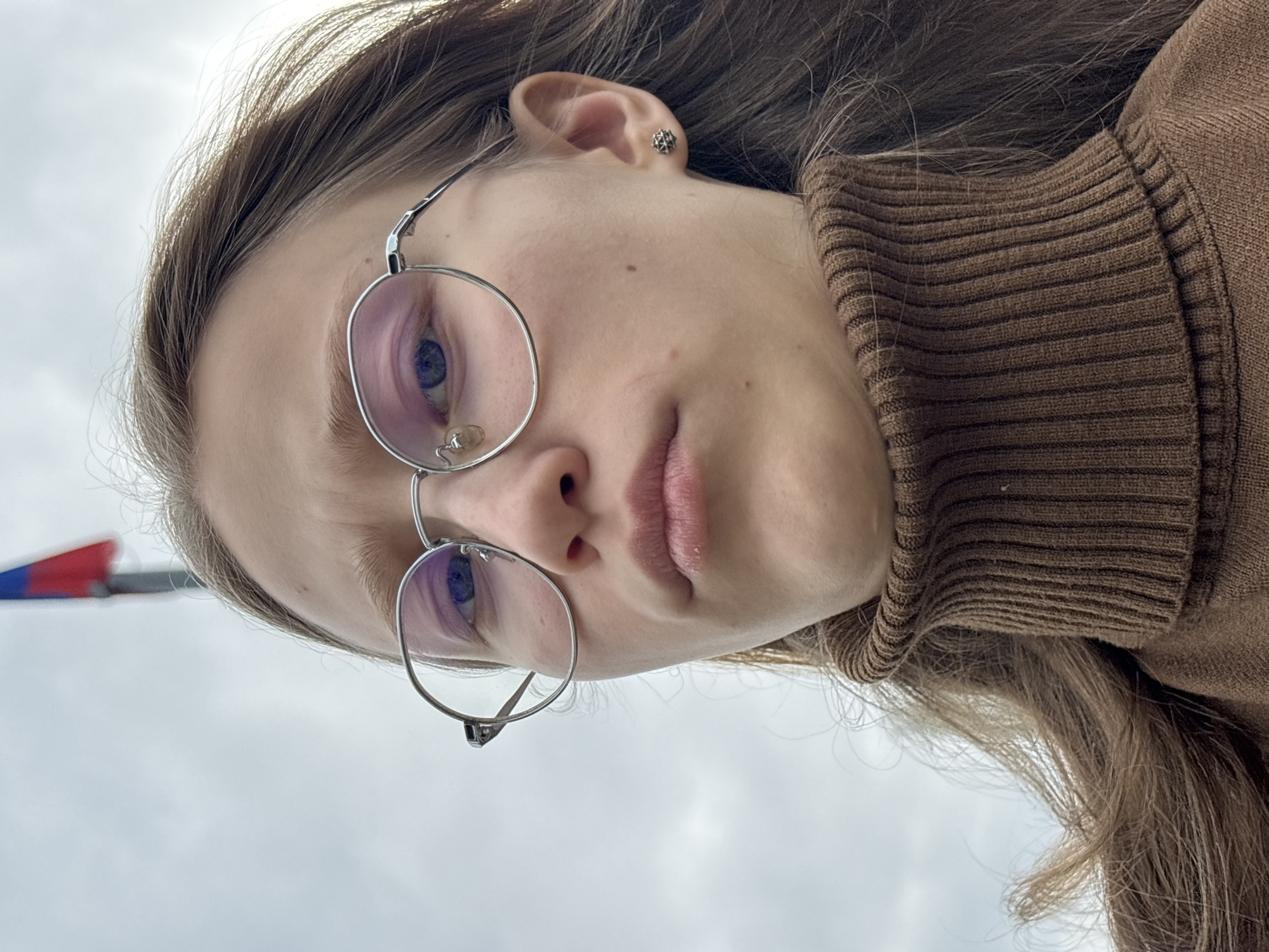 A young Caucasian woman with light brown hair, wearing round, silver-rimmed glasses and a brown turtleneck sweater, is centered in the frame. Her gaze is directed slightly downward, with a neutral expression. She has a small, geometric earring in her left ear. The background is a light gray sky, suggesting an outdoor setting. In the periphery, at the edge of the frame, a section of a red, white, and blue object is visible. This image was taken in Perm, Russia.