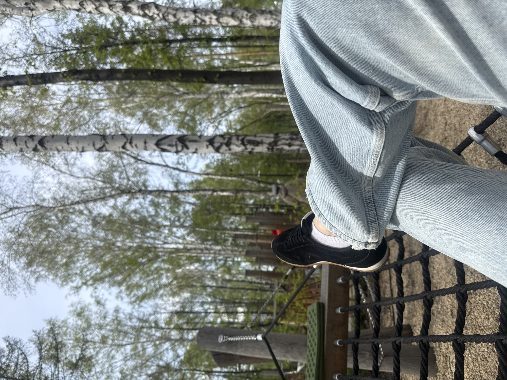 A person is sitting on a rope bridge in a park. The person is wearing light blue jeans, white socks, and black sneakers with a brown sole. The person's legs are crossed. In the background, there are birch trees, suggesting a park or forest setting. The photo was taken during daylight, with soft lighting and a slightly overcast sky visible through the trees. Given the reference to Yekaterinburg, Russia, the image could be taken in one of its many parks, such as the Central Park of Culture and Rest named after V.V. Mayakovsky or another similar outdoor recreational area with rope courses or adventure parks. The rope bridge itself appears to be part of a recreational structure or course.