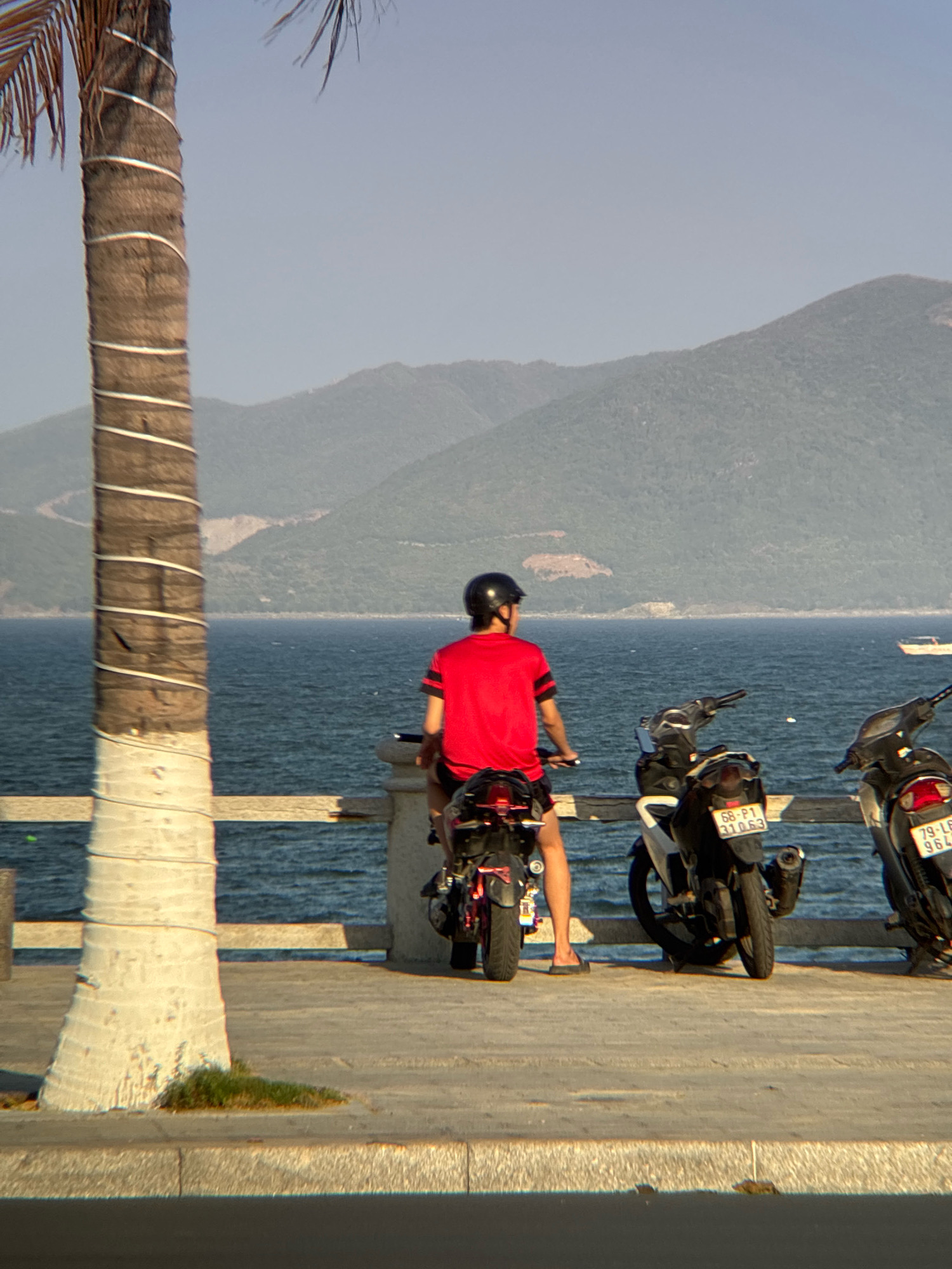 A person wearing a red shirt and black helmet is seated on a motorcycle, facing away from the camera. The motorcycle has a black frame and red accents. In the background, two other motorcycles are parked. One is black with white accents, and the other is black with red accents. The scene takes place on a paved walkway next to a body of water. In the distance, mountains are visible. The overall atmosphere suggests a coastal location, likely in Nha Trang, Vietnam, given the presence of palm trees, motorcycles, and the coastal mountain scenery.