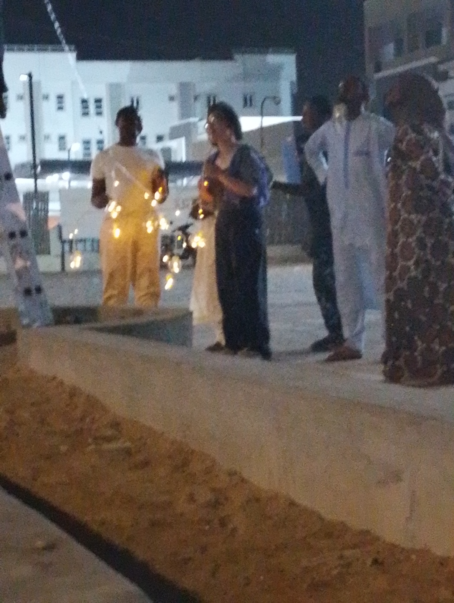 This nighttime image captures a gathering of several individuals on a paved surface in what appears to be Gandu Albasa, Nigeria. The dark sky indicates the scene is taking place after sunset, with artificial light sources providing illumination.
In the center-left, a person is prominently featured, wearing a white t-shirt and light-colored pants, extensively adorned with glowing string lights that wrap around their torso and legs. To their immediate right, another individual, dressed in dark clothing, also appears to be decorated with similar string lights, casting a warm, golden glow. These lights suggest a festive or celebratory event.
Further to the right, a man stands in a light blue traditional garment, likely a thobe or jalabiya, topped with a dark cap. He holds a relaxed posture with his hands on his hips. Next to him, on the far right, a person is partially visible, wearing a long, patterned, dark-colored dress or robe. On the far left edge of the frame, another individual in a light-colored garment is partially visible, next to a ladder-like metallic structure.
The background consists of several modern, white multi-story buildings with numerous windows, suggesting an urban or residential area. A streetlight or similar fixture is visible amidst the buildings. The ground in the foreground is a paved surface, with a slightly raised curb or divider running diagonally across the lower portion of the frame. The overall image has a somewhat blurry quality, typical of low-light photography. There is no visible text in the image.